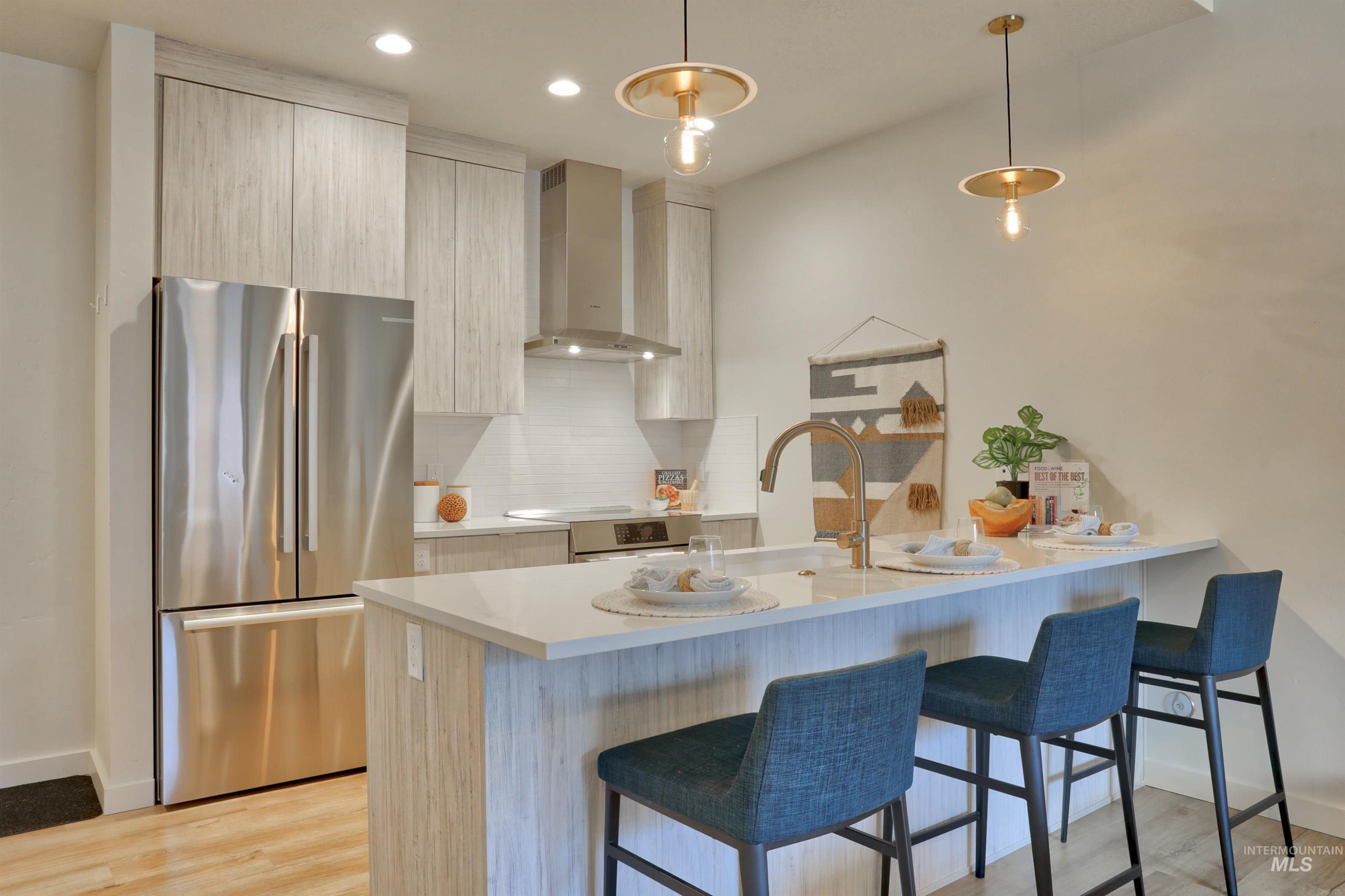 Kitchen with freestanding refrigerator, wall chimney range hood, modern cabinets, backsplash, and decorative light fixtures