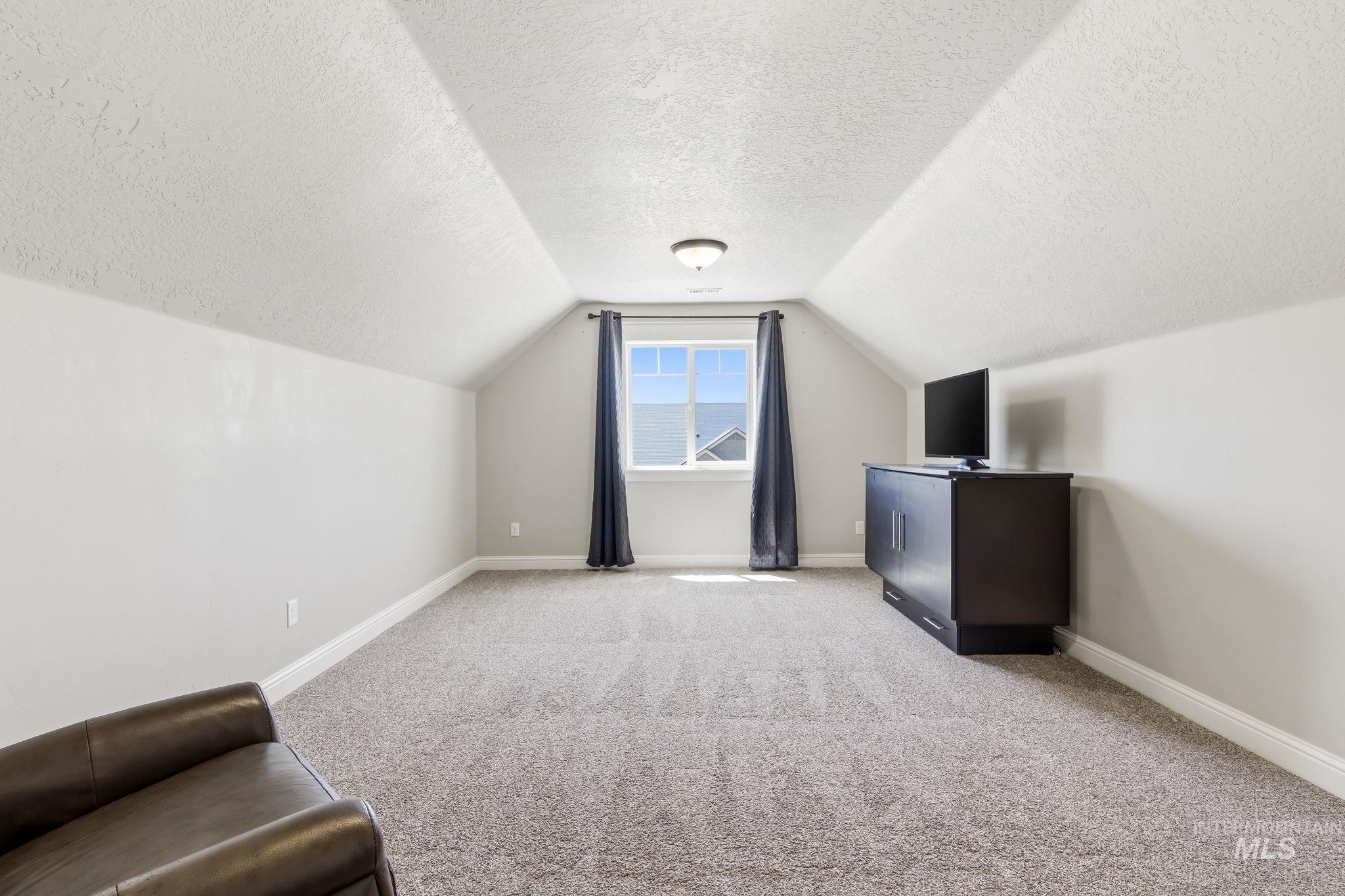 Spacious carpeted room featuring a window with dark drapes, sloped ceilings, and white baseboards