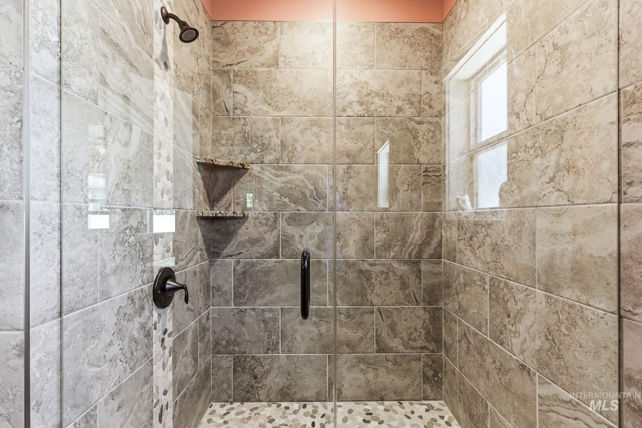 Walk-in shower with full height stone-finish tile, oil-rubbed bronze fixtures, a frameless glass enclosure, built-in shelving, and a window