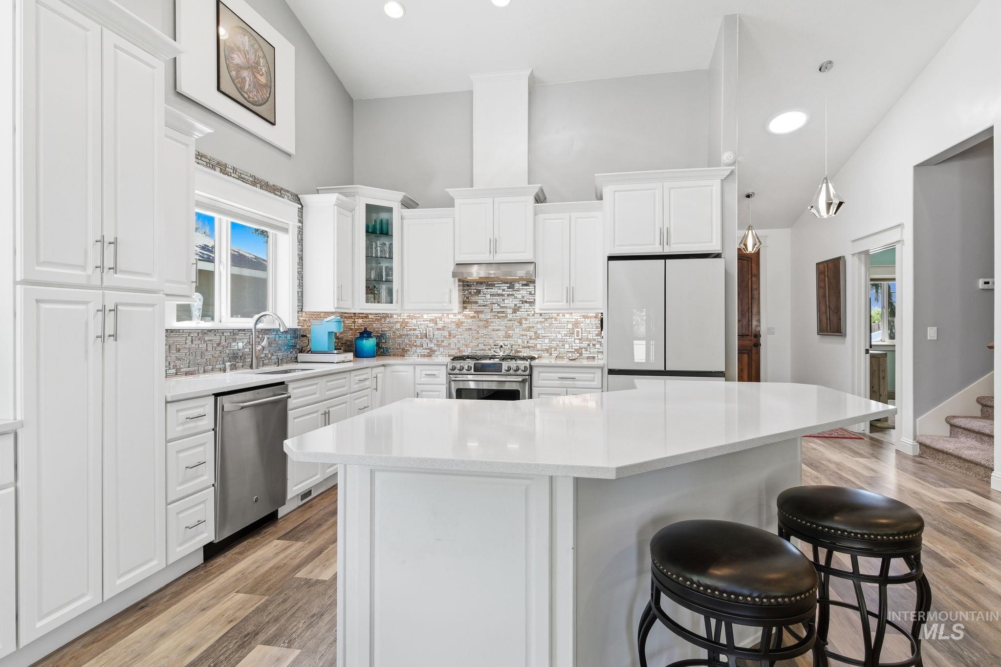 Open-concept kitchen featuring vaulted ceilings, white shaker cabinetry, stainless steel appliances, a multi-toned tile backsplash, and a central island with a solid surface countertop