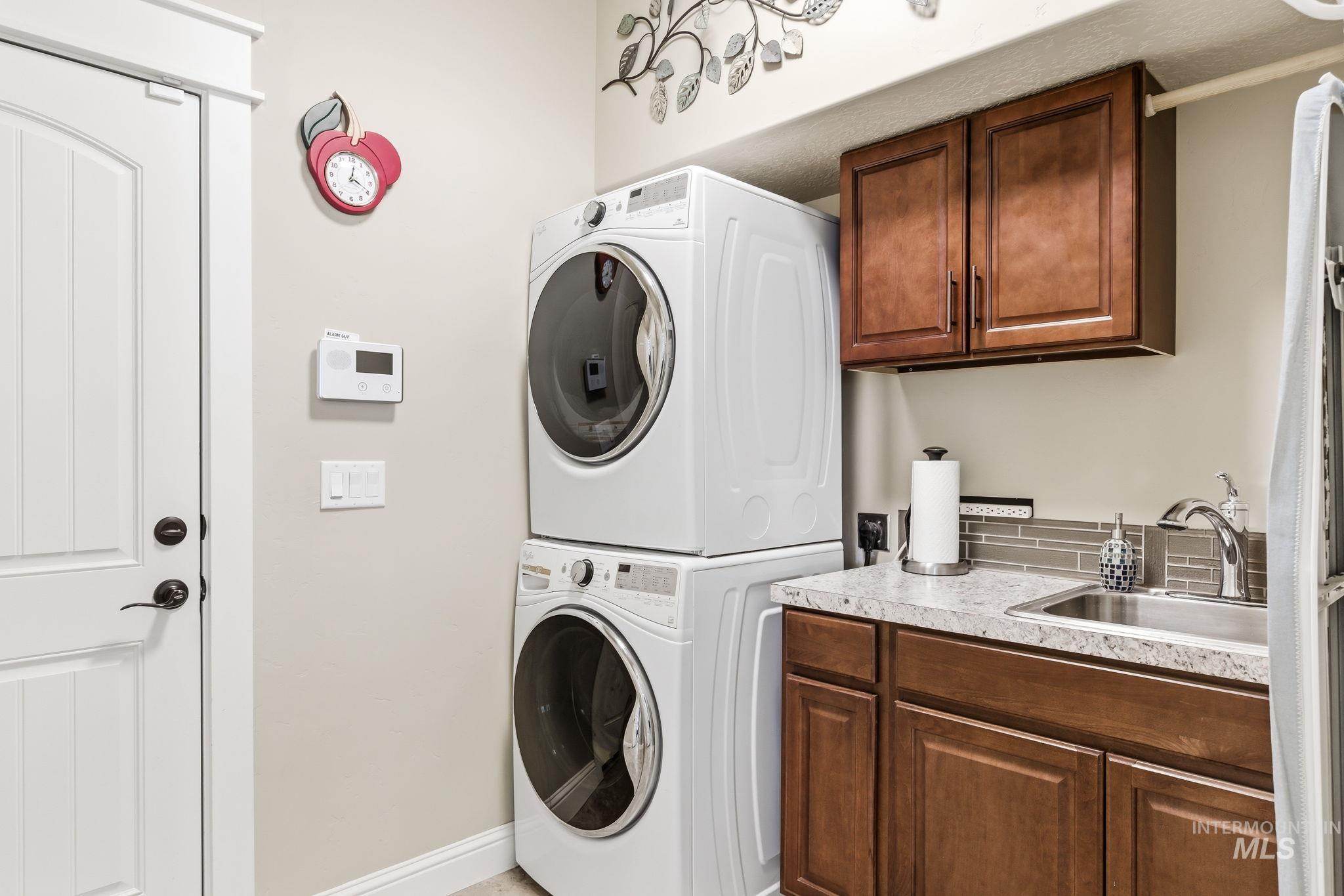 Dedicated laundry area featuring stacked appliances, a utility sink, dark wood-finish cabinetry, light-toned countertop, and a gray subway tile backsplash