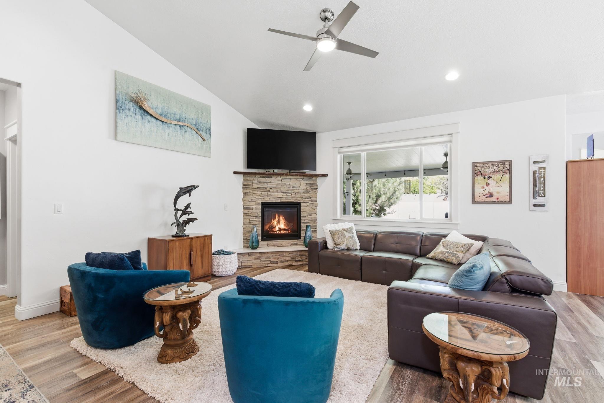 Spacious living area featuring vaulted ceilings, a corner stone-faced fireplace, a large picture window, a contemporary ceiling fan, and wood-finish flooring