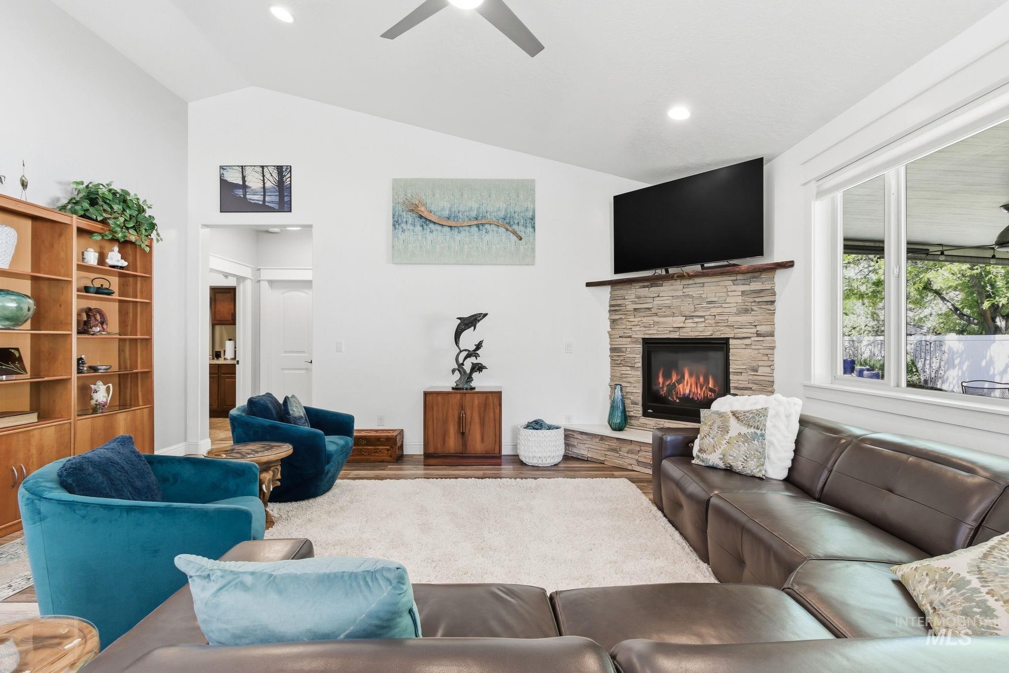 Vaulted ceilings complement the living area featuring a stacked stone fireplace with wood mantel, large windows, and a ceiling fan