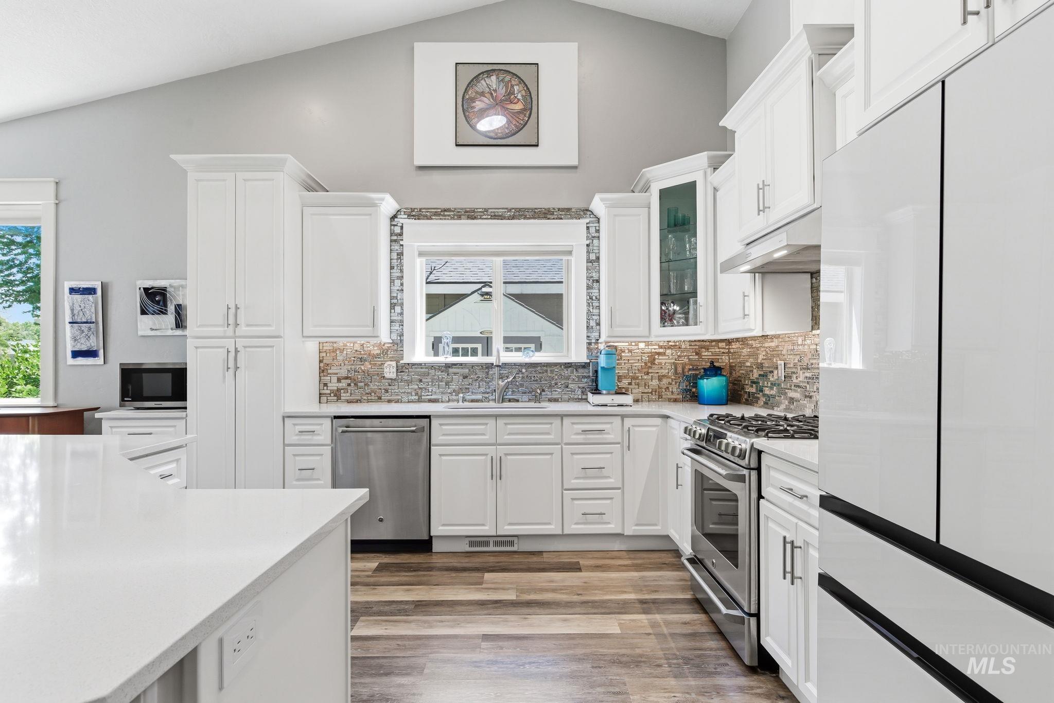 Kitchen featuring white cabinetry, wood-finish flooring, stainless steel appliances, a multi-tone tile backsplash, and a vaulted ceiling