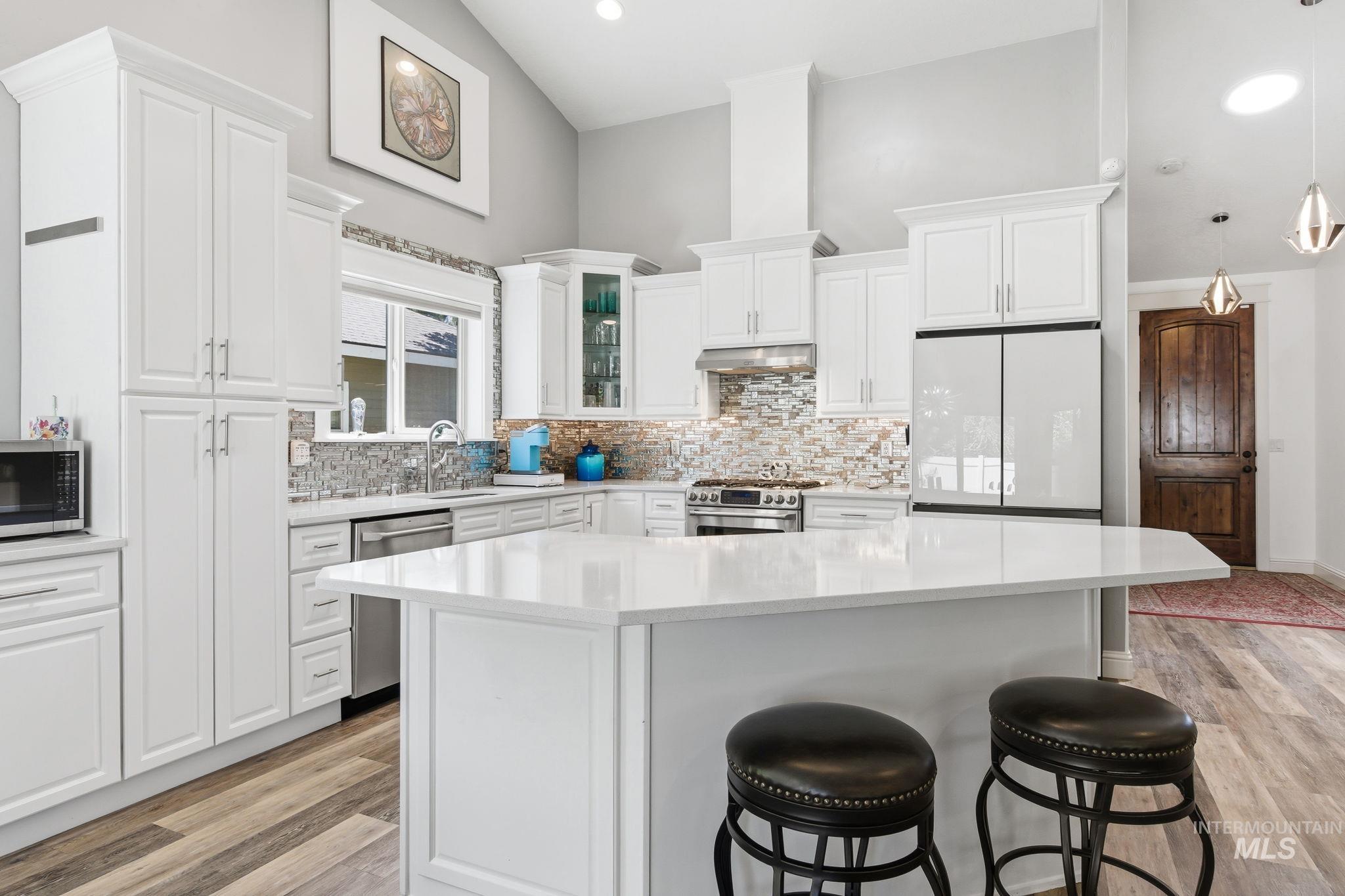 Modern kitchen featuring a central island with a light-toned countertop, white cabinetry, a mosaic tile backsplash, stainless steel appliances, and wood-finish flooring