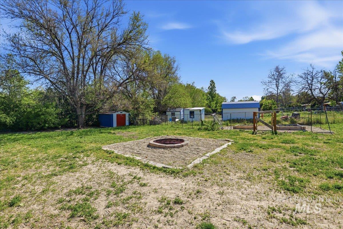 Expansive backyard featuring a central fire pit area with paver edging, a large mature tree, two storage sheds, raised garden beds with a wood gate, and perimeter fencing