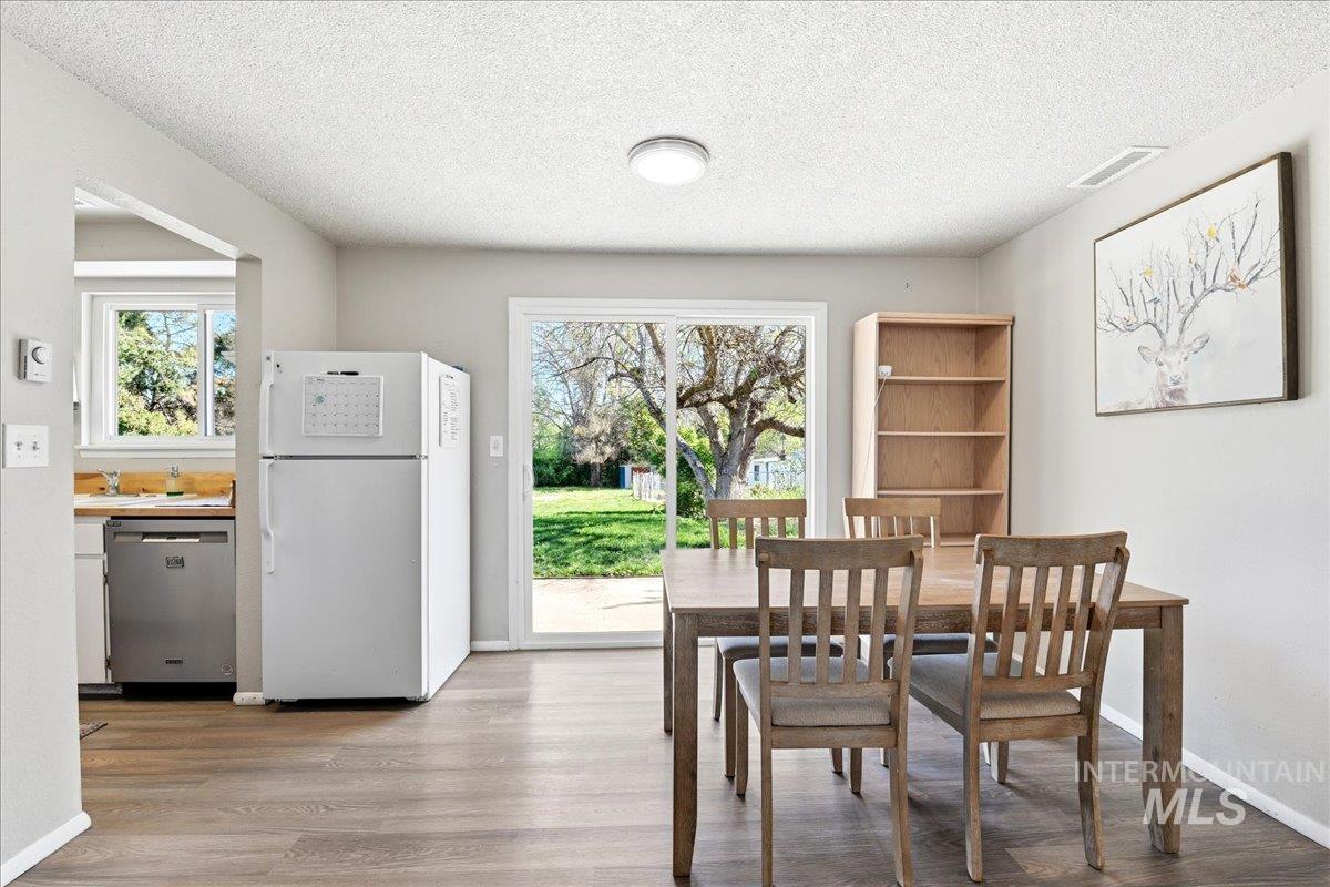 Open dining area with wood-finish flooring, featuring a sliding glass door to the backyard, a ceiling-mounted light fixture, and an integrated kitchen space with a white refrigerator and a stainless steel dishwasher