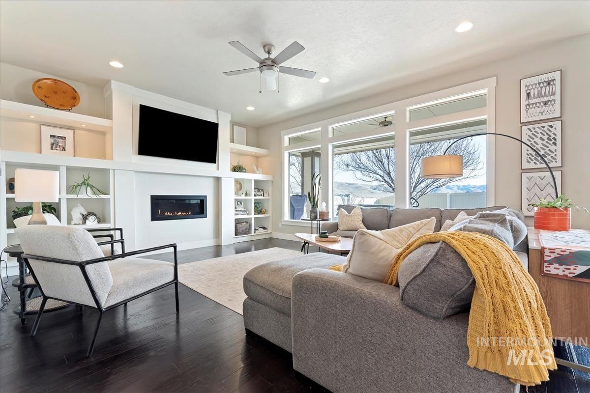 Living room with built in shelves, a glass covered fireplace, dark wood finished floors, ceiling fan, and recessed lighting