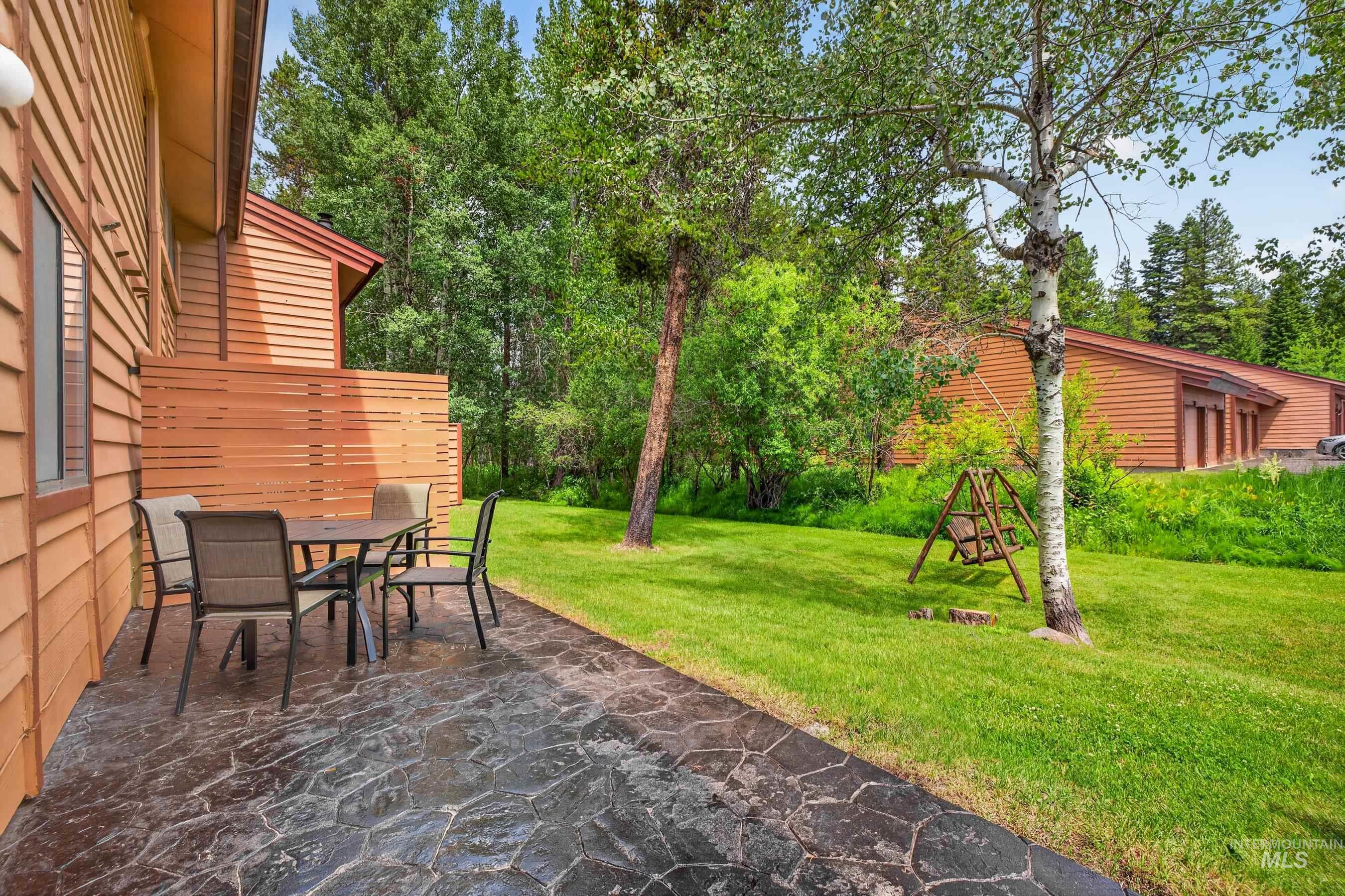 Exterior patio featuring a textured stone-finish surface, slatted privacy fence, and a manicured lawn surrounded by mature trees