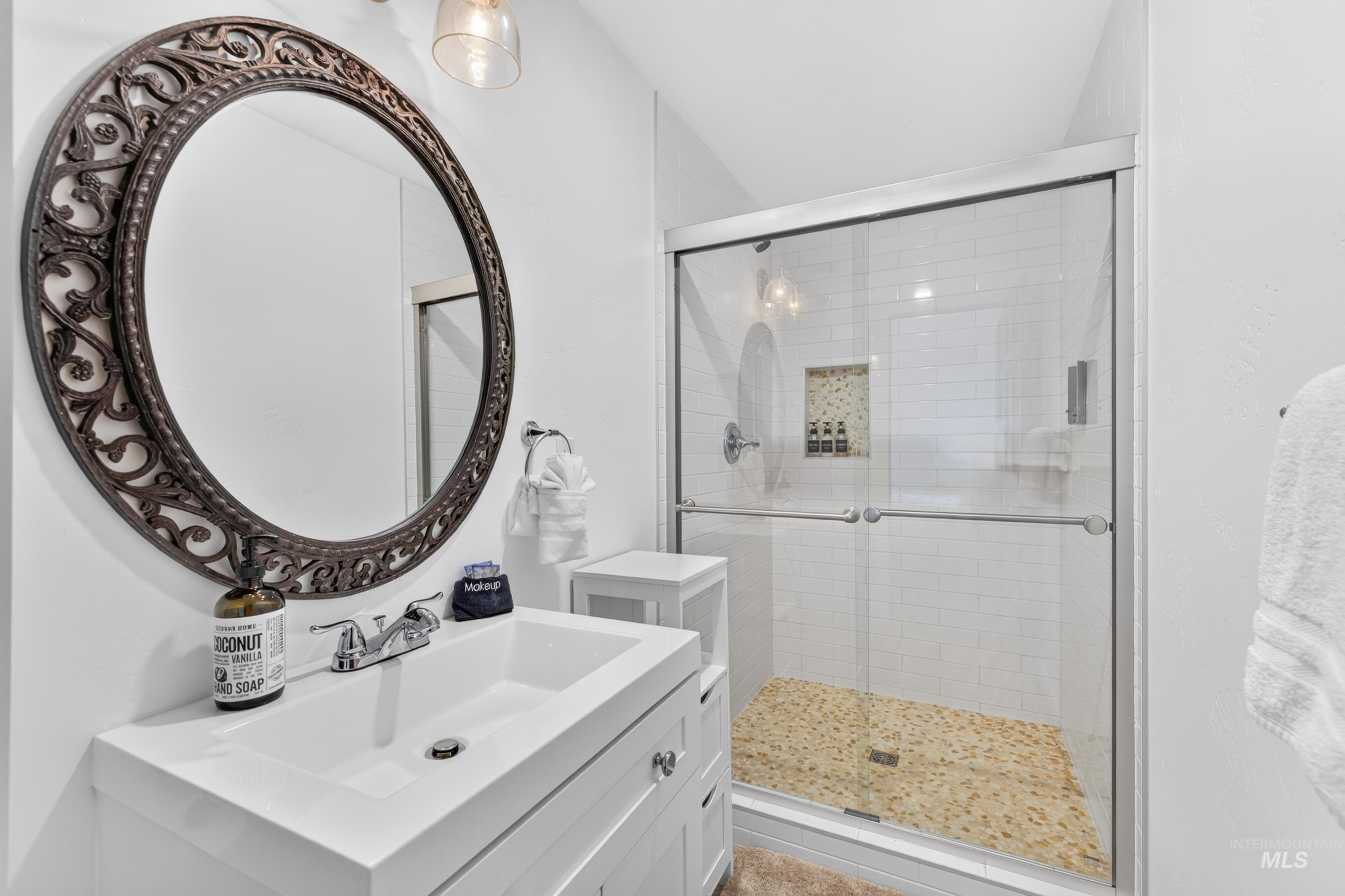 Bathroom featuring a white vanity with an integrated sink, a dark bronze ornate oval mirror, and a glass-enclosed shower with subway tile walls and a pebble-style floor