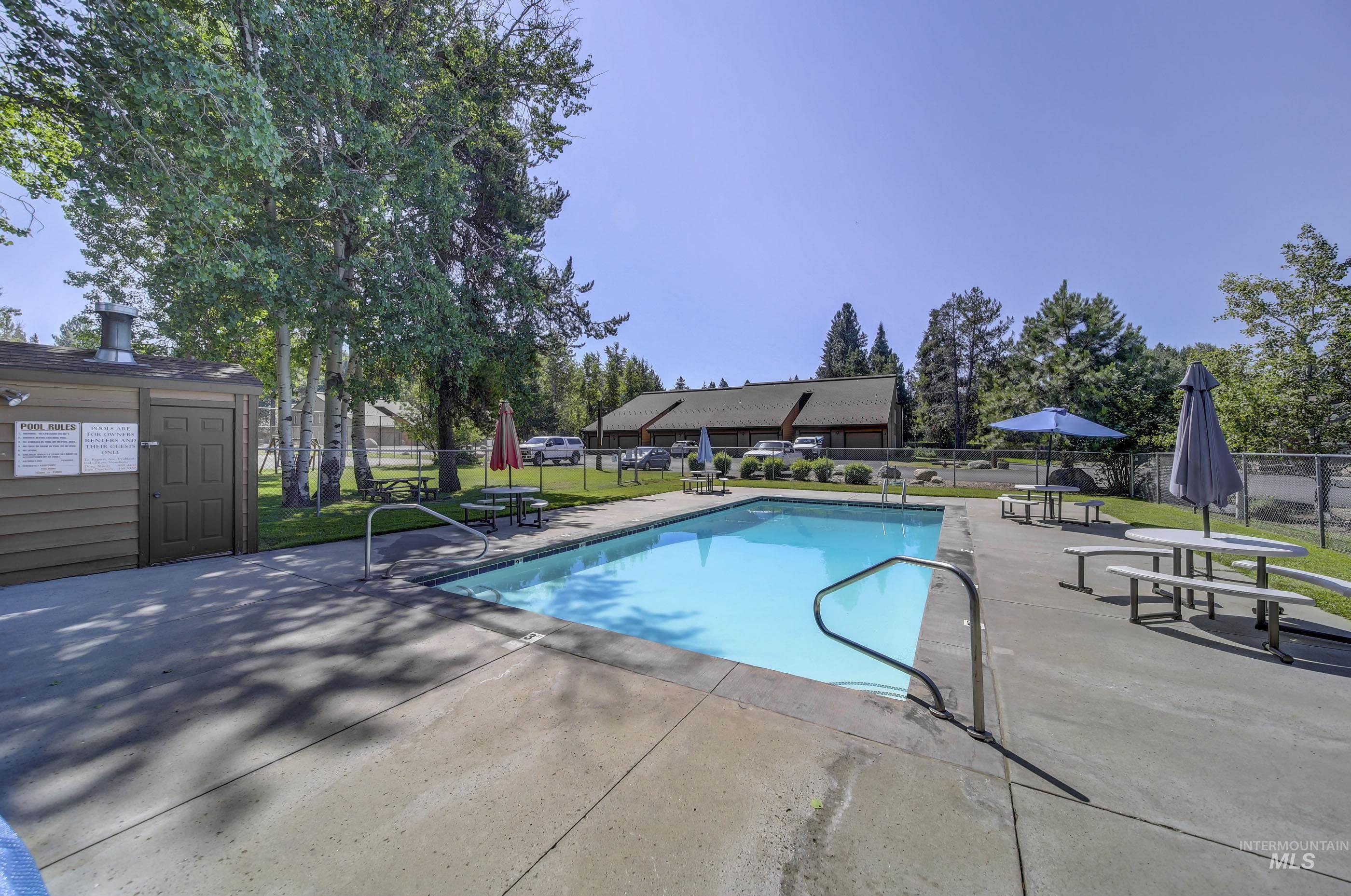 Outdoor swimming pool with concrete decking, surrounded by mature trees, a chain-link fence, and a utility building