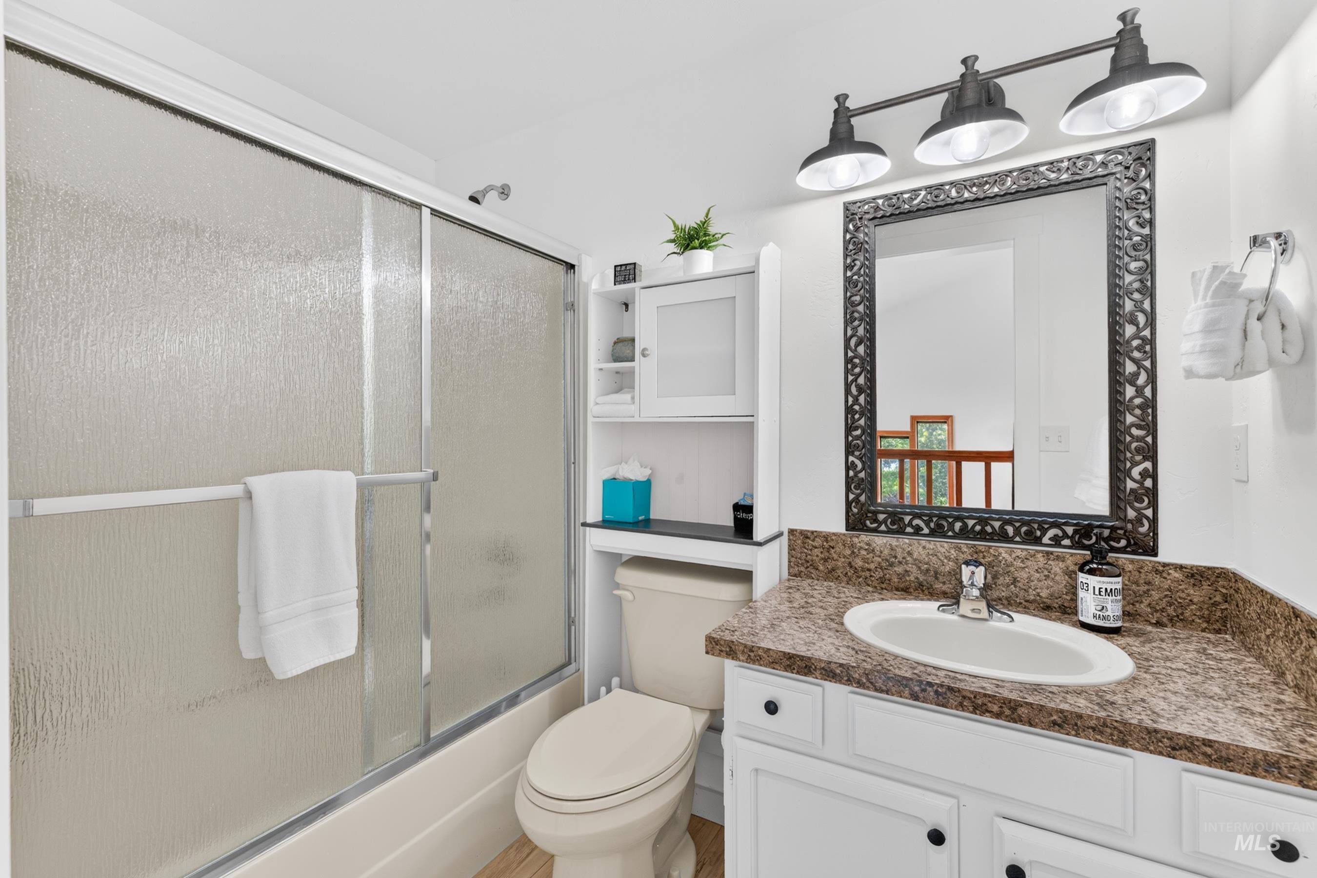 Bathroom featuring a tub with sliding frosted glass enclosure, white vanity with a light speckled countertop and integrated sink, decorative framed mirror, and a three-light fixture
