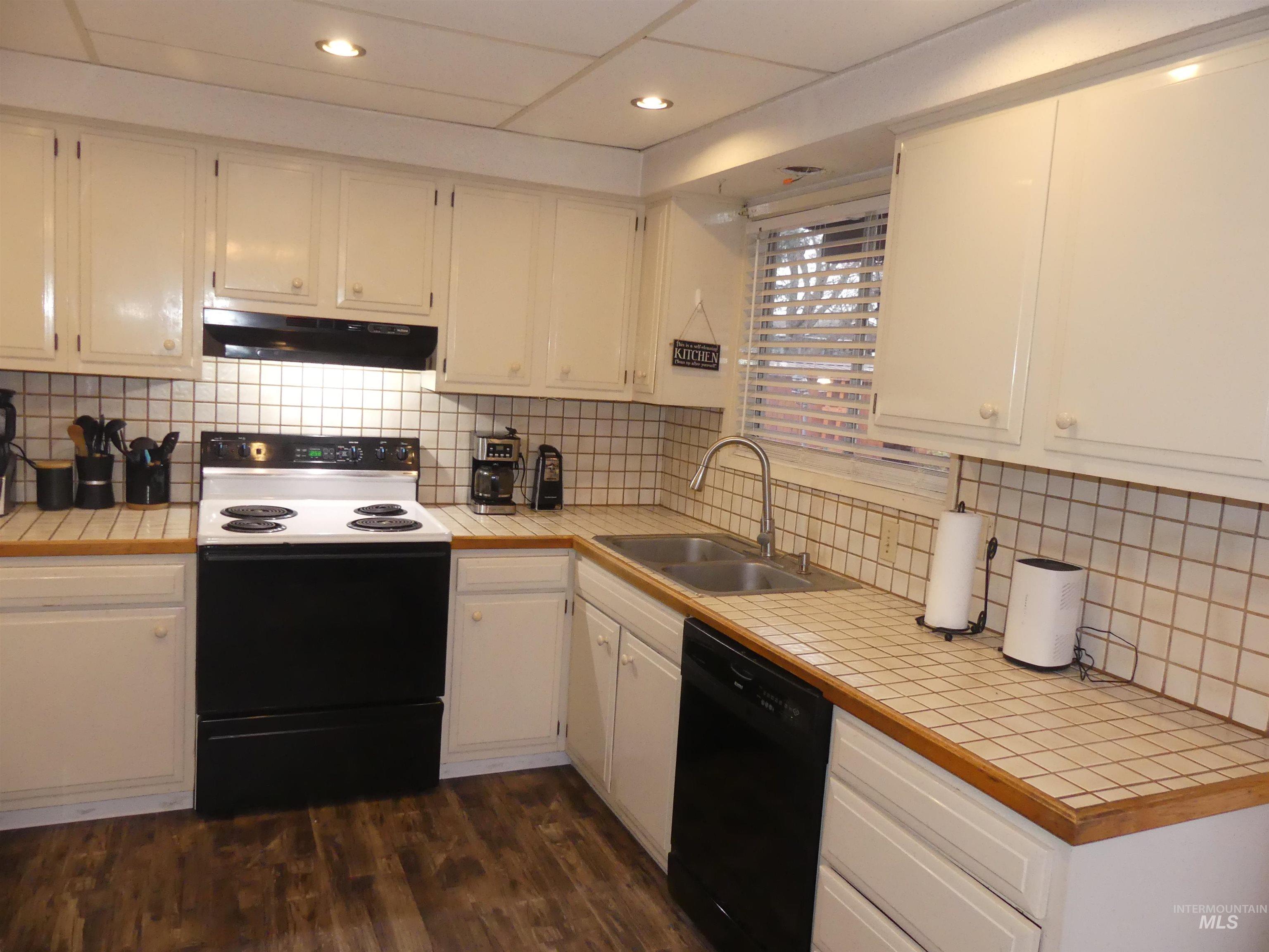 Kitchen with tile countertops, black appliances, tasteful backsplash, under cabinet range hood, and recessed lighting