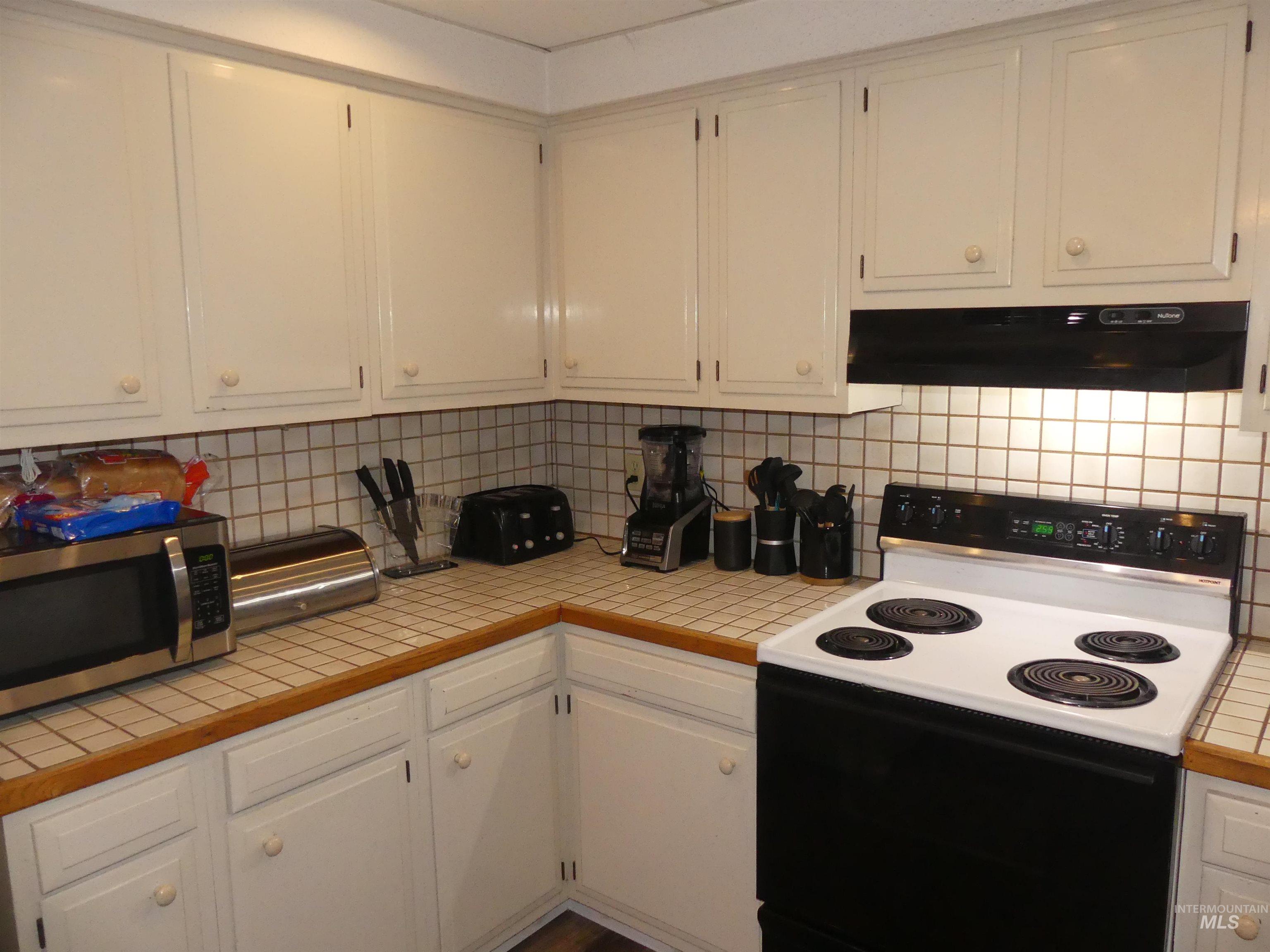 Kitchen with range with electric stovetop, tile countertops, stainless steel microwave, extractor fan, and white cabinetry