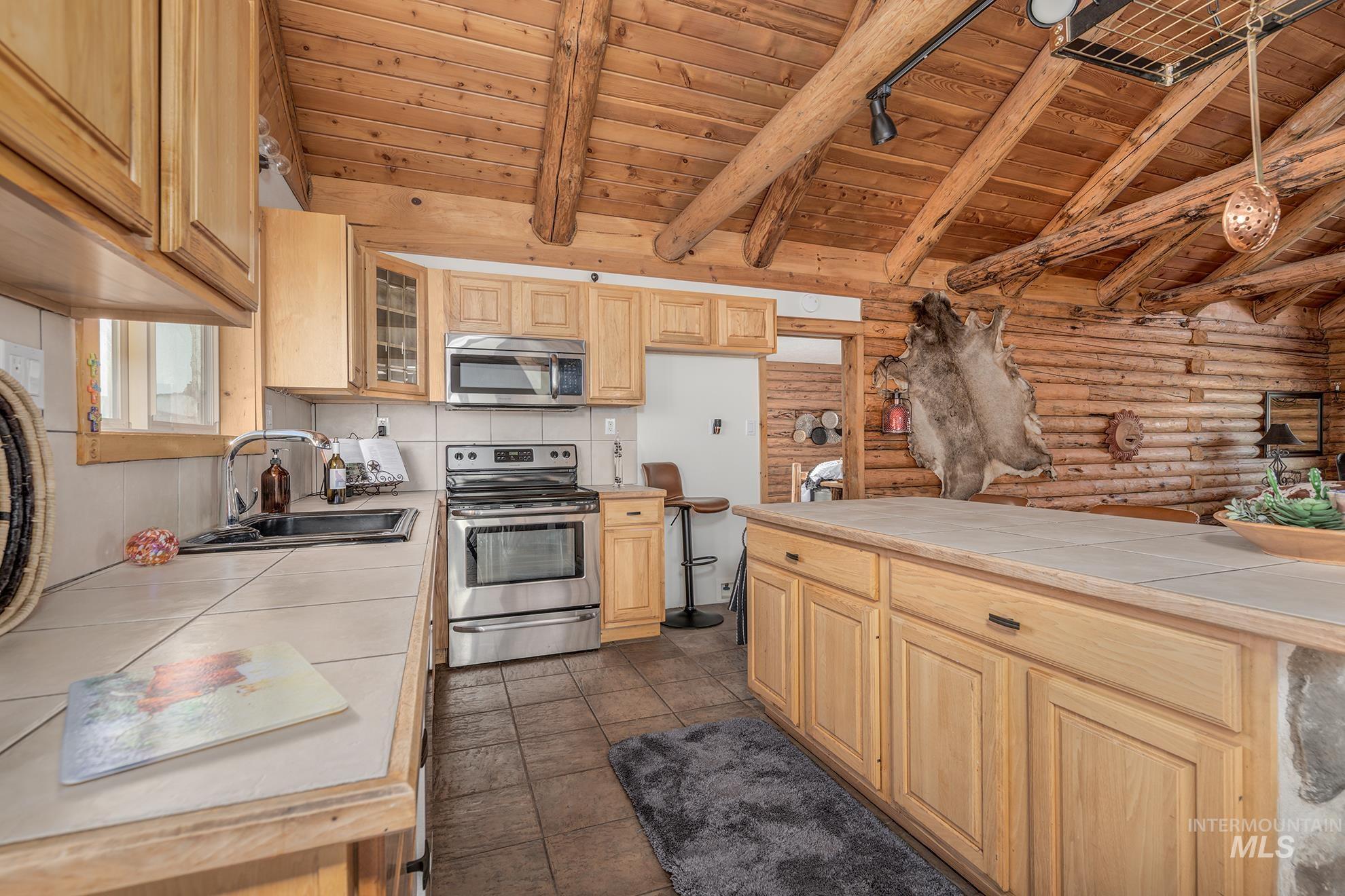 Kitchen with tile counters, wooden ceiling, log walls, stainless steel appliances, and glass fronted cabinets