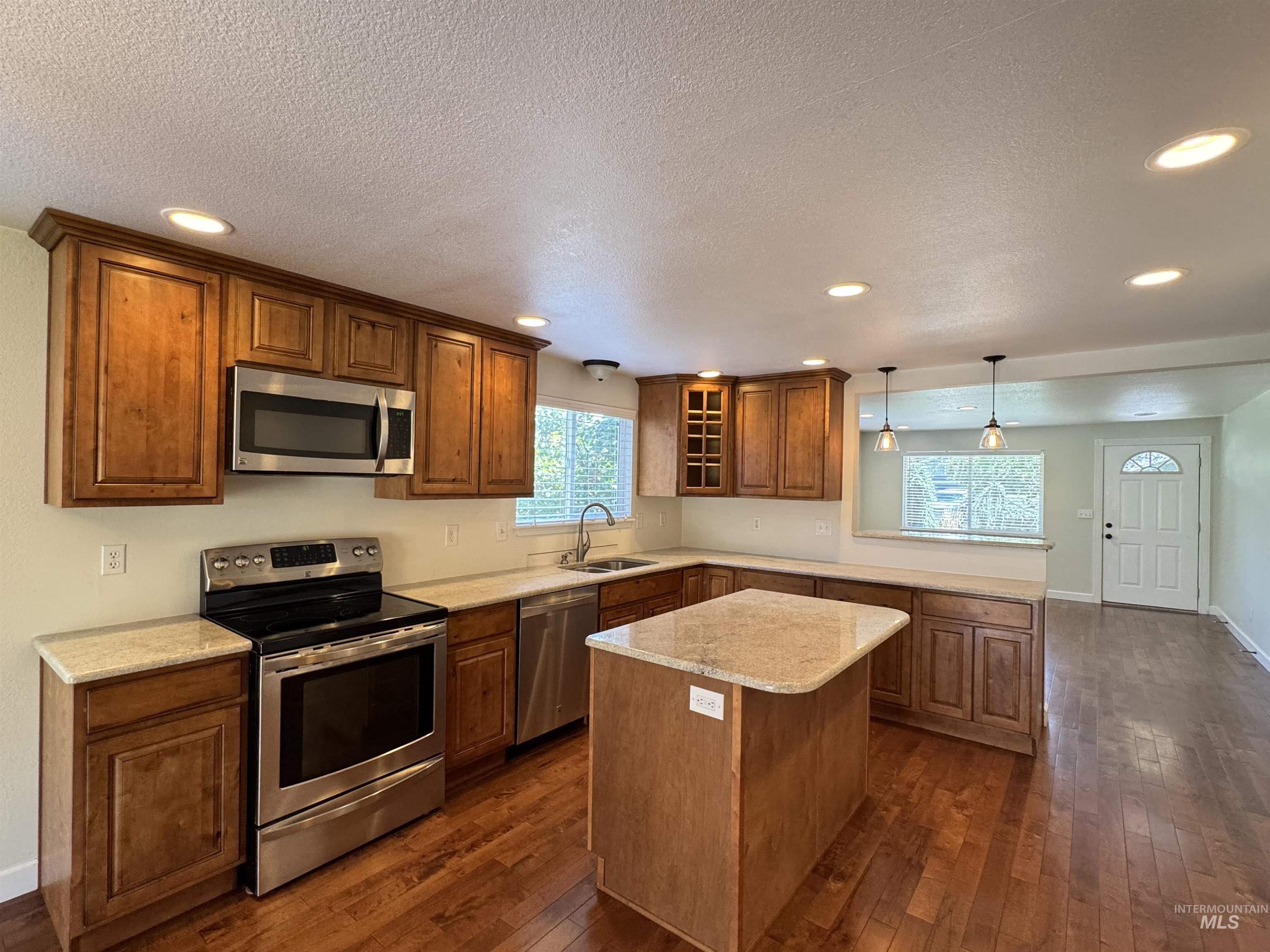 Kitchen featuring appliances with stainless steel finishes, a peninsula, dark wood-style flooring, brown cabinetry, and glass insert cabinets