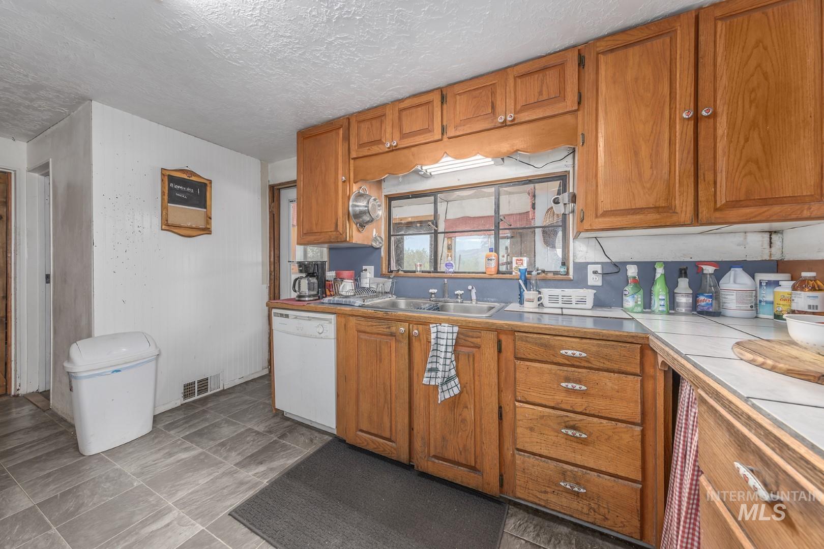 Kitchen with brown cabinetry, dishwasher, a textured ceiling, and tile countertops