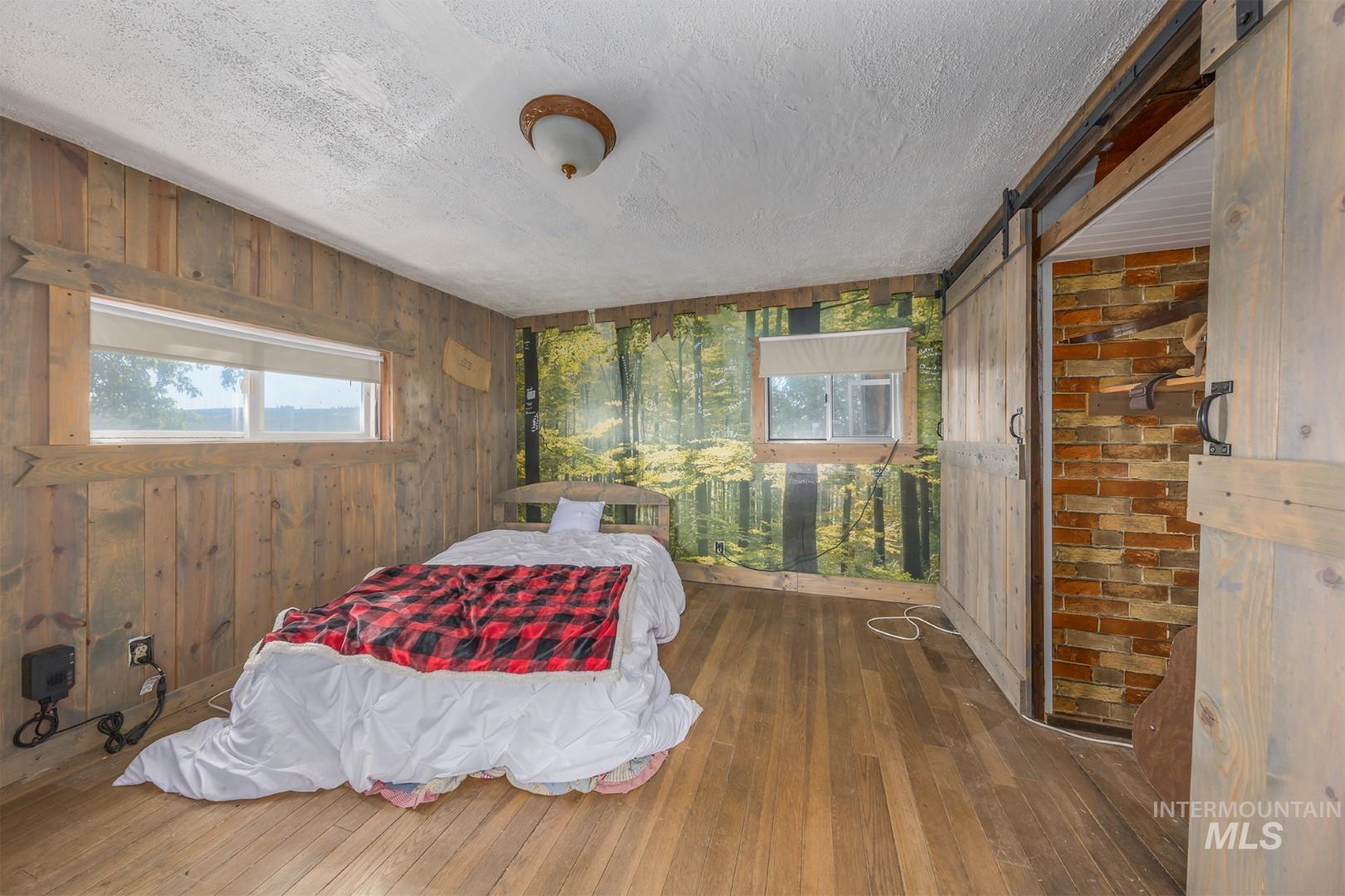 Bedroom with wooden walls, a barn door, hardwood / wood-style floors, a textured ceiling, and multiple windows