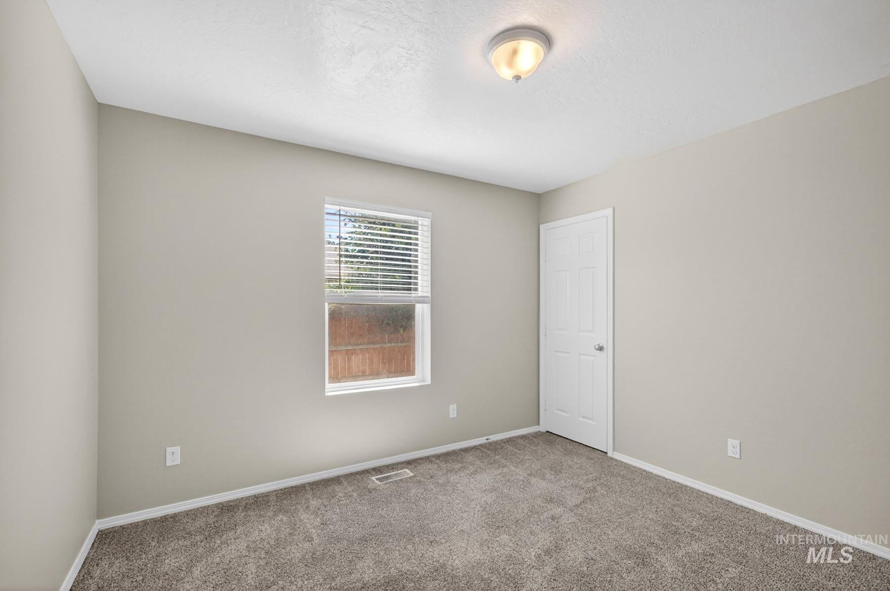Neutral-toned interior room featuring textured carpeting, a single window with horizontal blinds, and a paneled white door