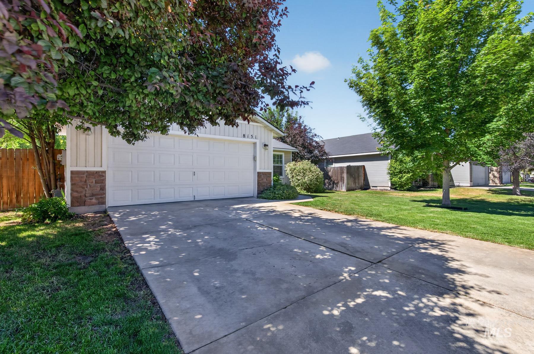 Front exterior featuring a white paneled garage door, stone facade accents, vertical white siding, a concrete driveway, and mature landscaping