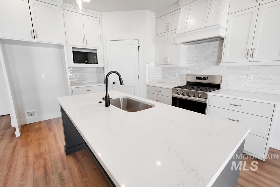 Kitchen featuring stainless steel range with gas stovetop, a kitchen island with sink, white cabinets, light wood finished floors, and decorative backsplash