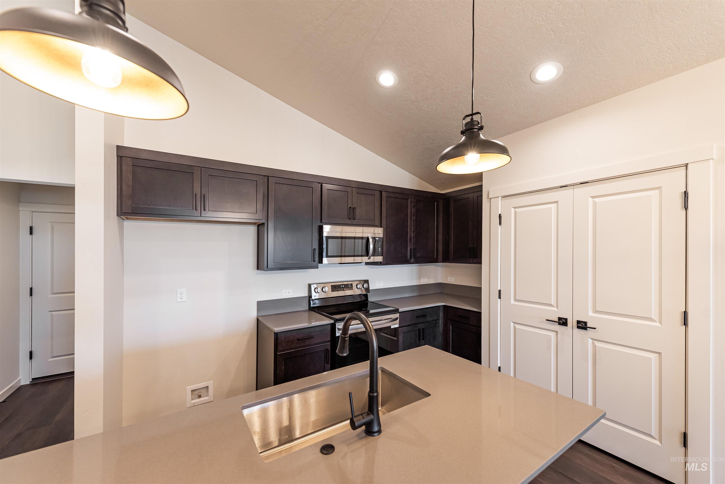 Kitchen featuring dark brown cabinets, appliances with stainless steel finishes, dark wood-style floors, lofted ceiling, and hanging light fixtures