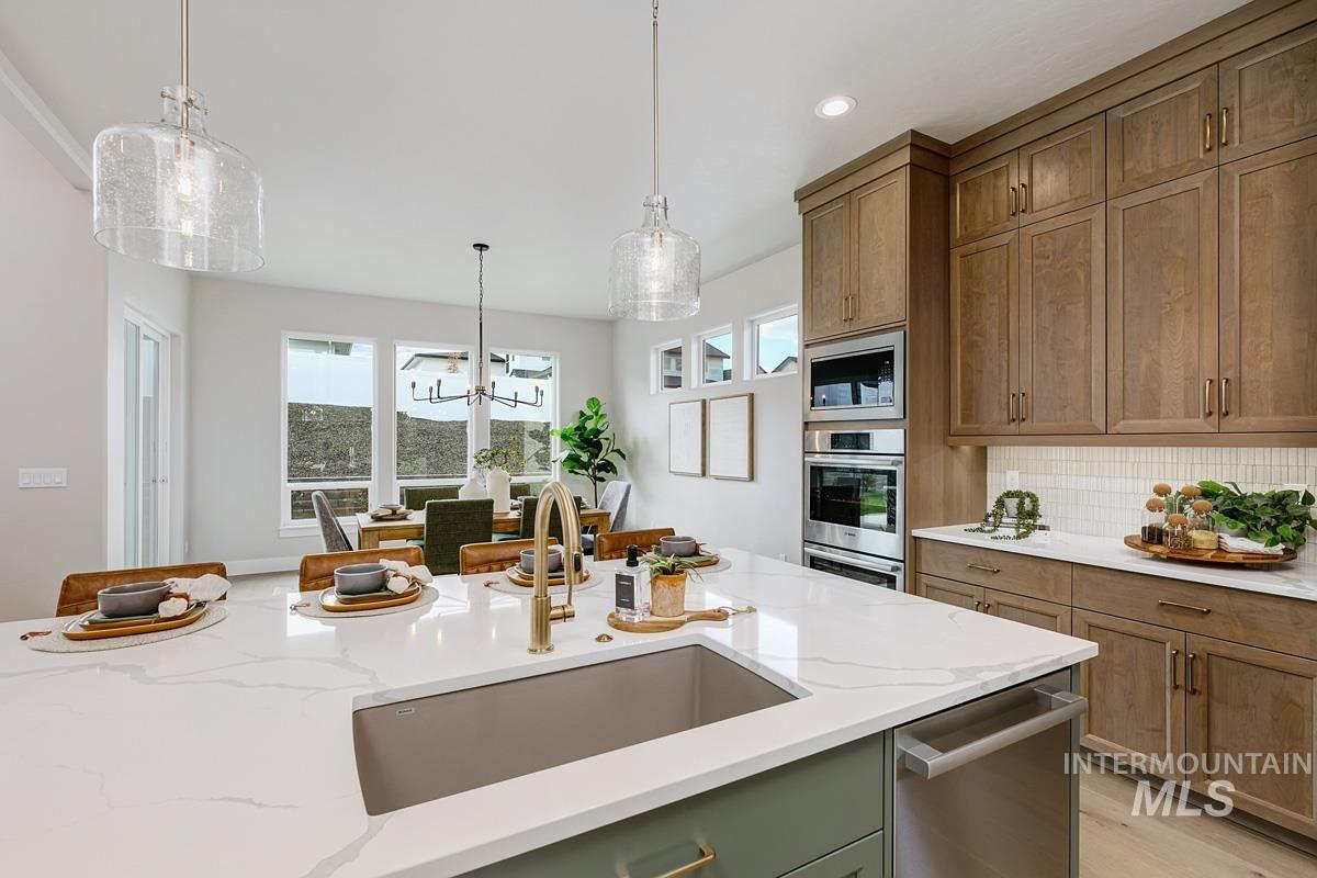 Kitchen featuring light stone counters, hanging light fixtures, tasteful backsplash, brown cabinets, and appliances with stainless steel finishes