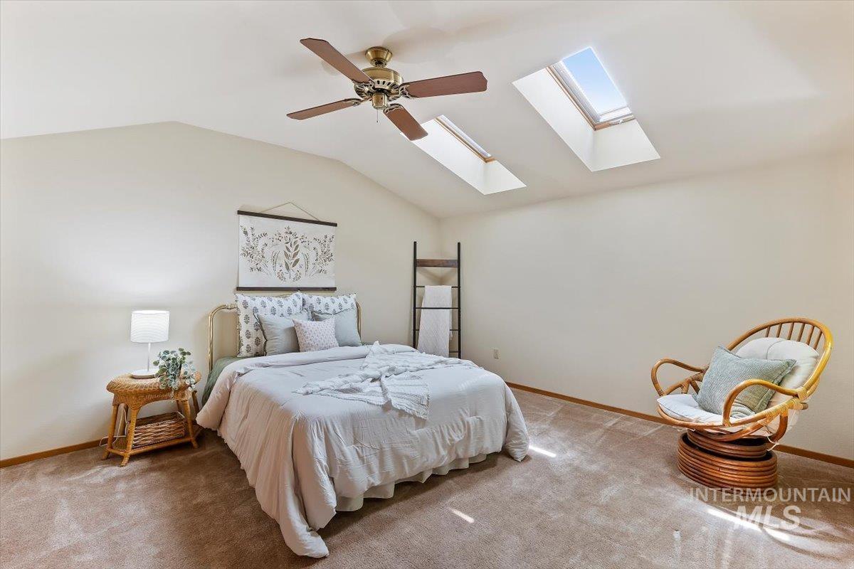 Vaulted ceiling room featuring two skylights, a ceiling fan, light beige walls, and brown carpet flooring