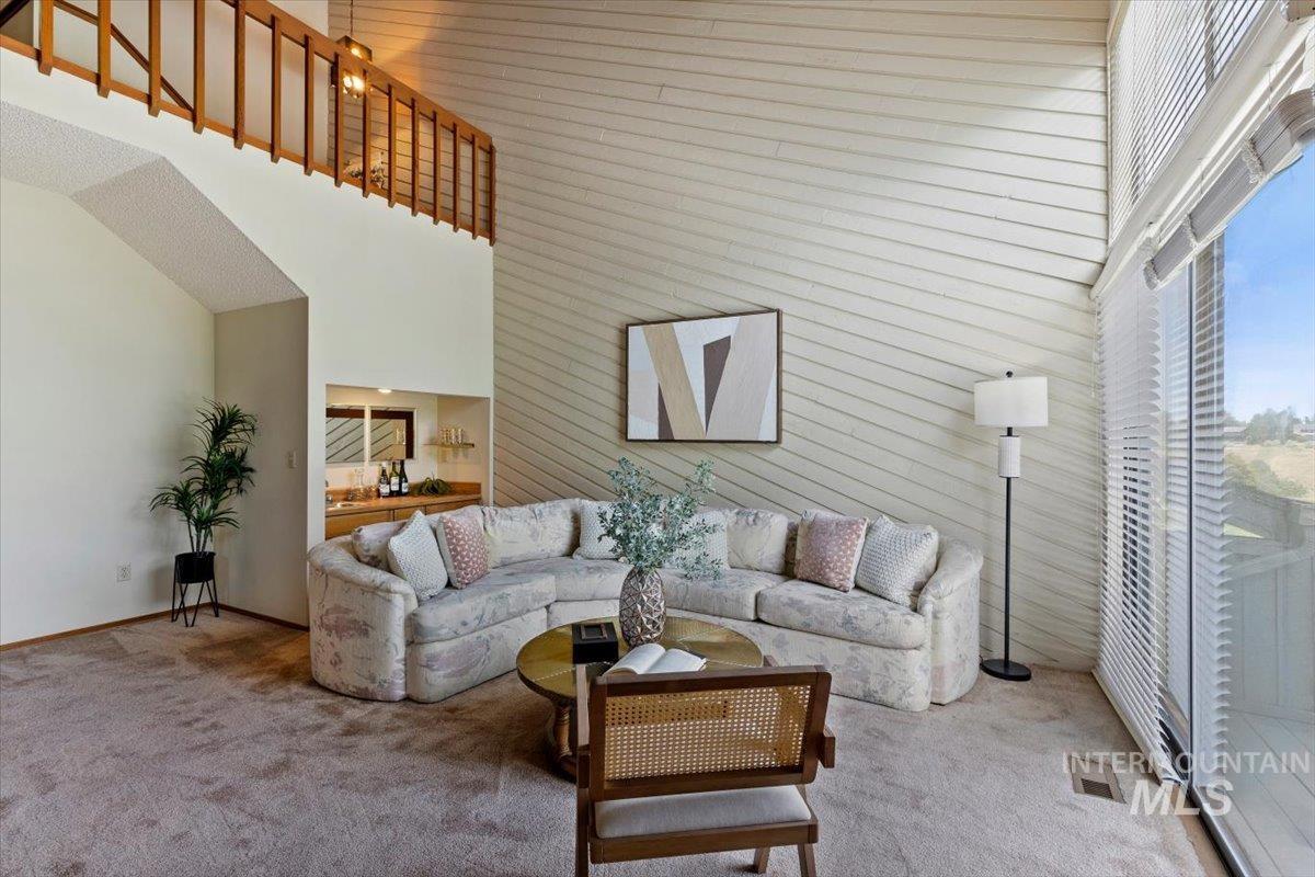 Living area featuring high ceilings, a built-in wet bar with mirror, a second-story balcony with wood railing, large windows with blinds, and wall paneling