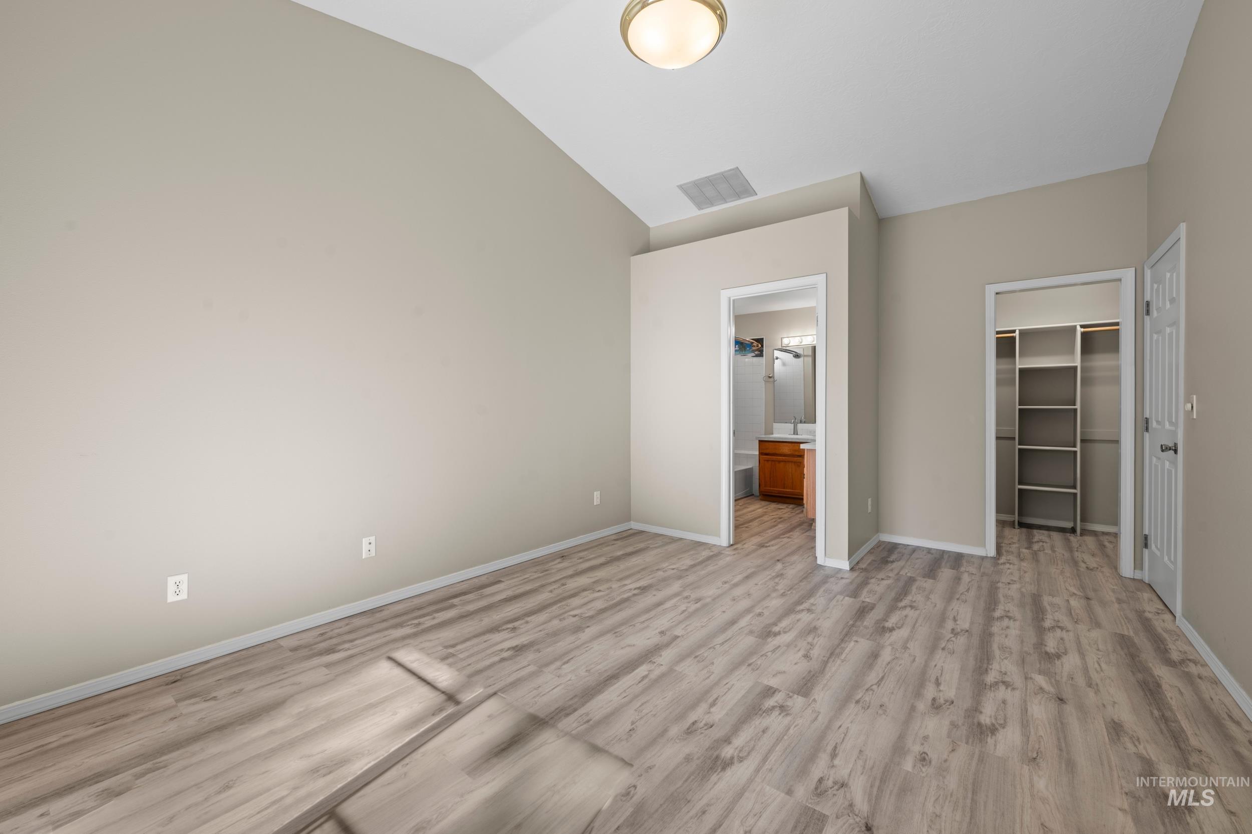 Unfurnished bedroom featuring a spacious closet, ensuite bathroom, light wood finished floors, and vaulted ceiling