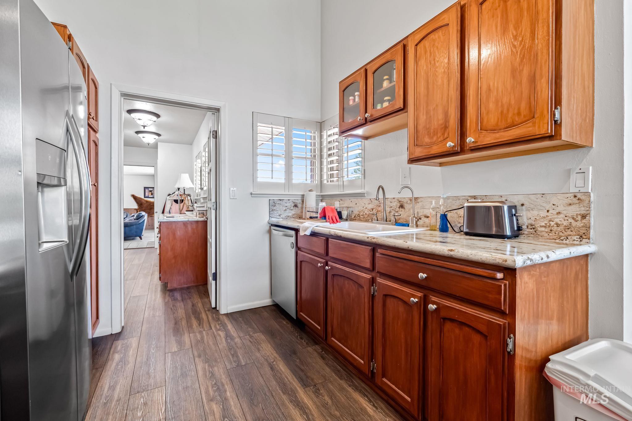 Kitchen featuring appliances with stainless steel finishes, dark wood-type flooring, glass insert cabinets, brown cabinets, and light stone counters