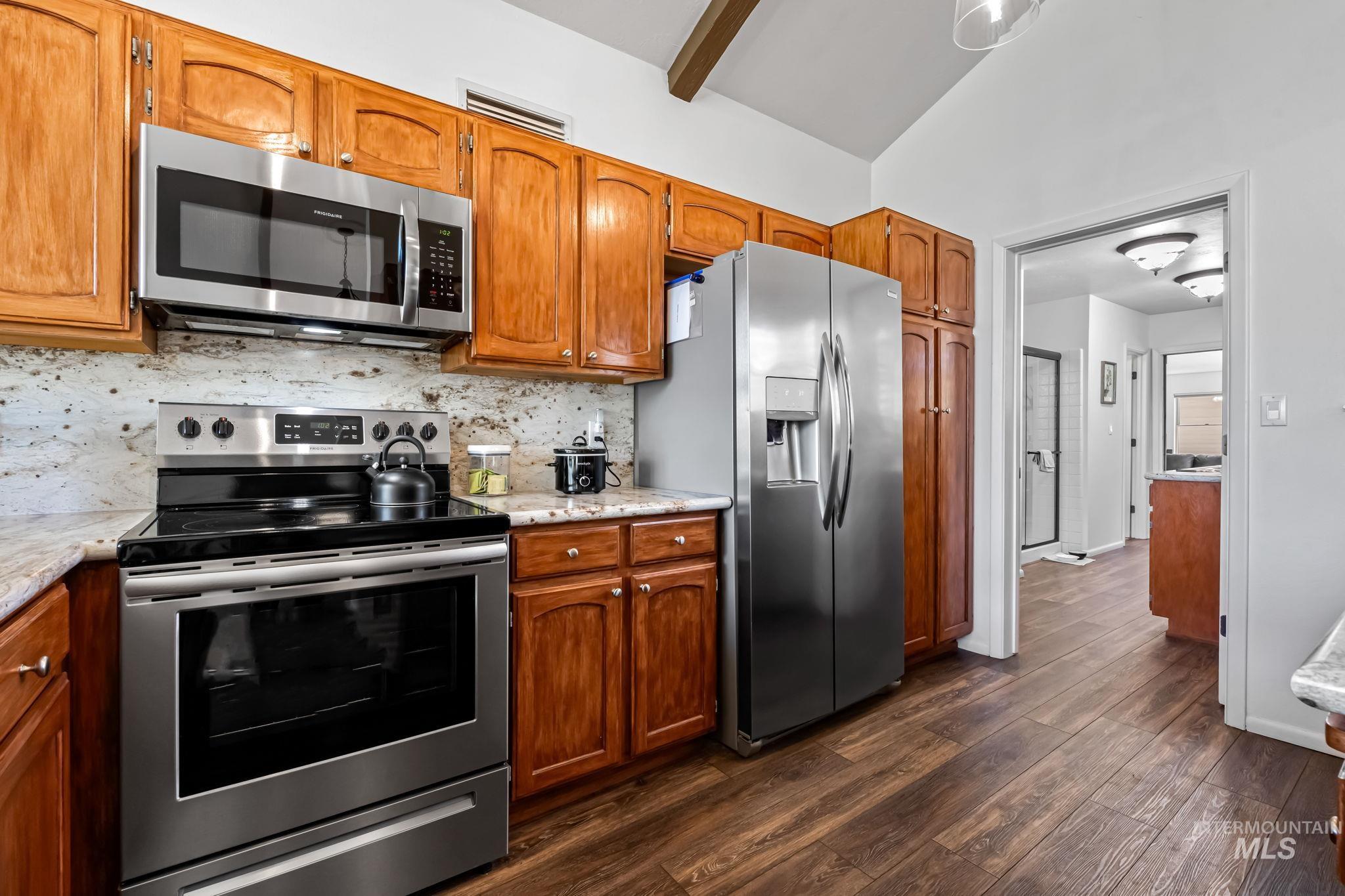 Kitchen featuring stainless steel appliances, backsplash, brown cabinetry, dark wood-style flooring, and lofted ceiling