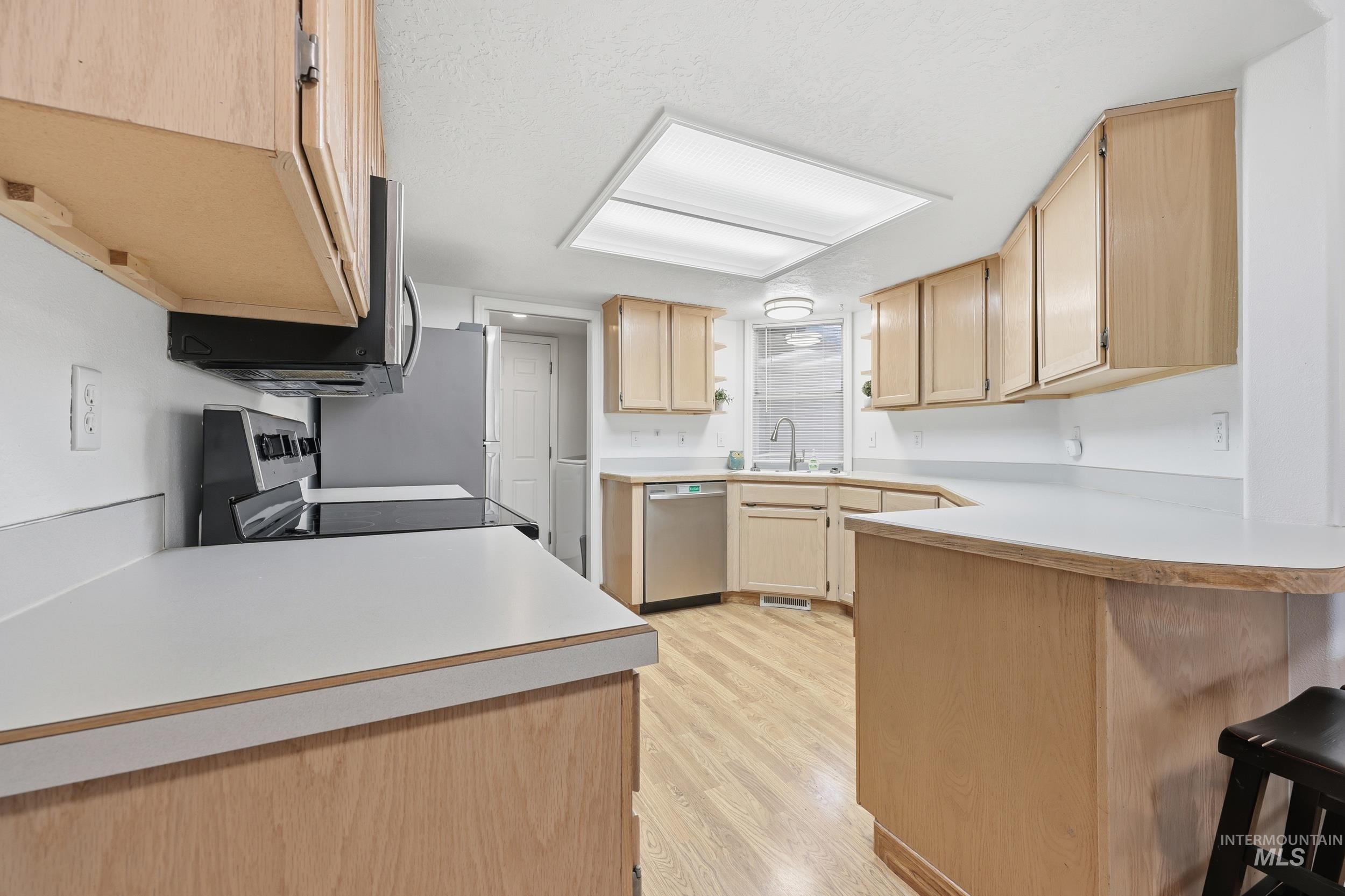 Kitchen with light brown cabinetry, light countertops, a peninsula, appliances with stainless steel finishes, and light wood-style flooring