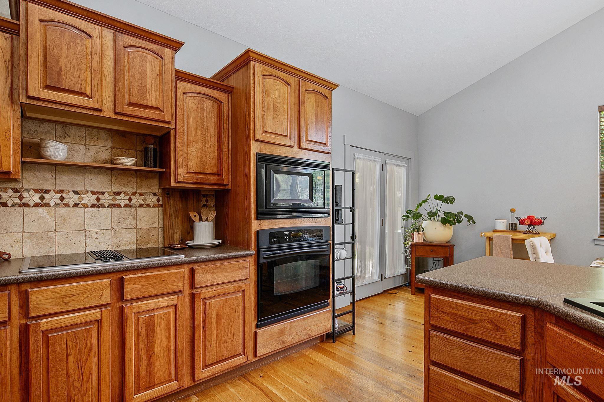 Kitchen featuring wood-finish cabinetry, a built-in microwave and oven, a cooktop, a tile backsplash with decorative accents, and hardwood flooring