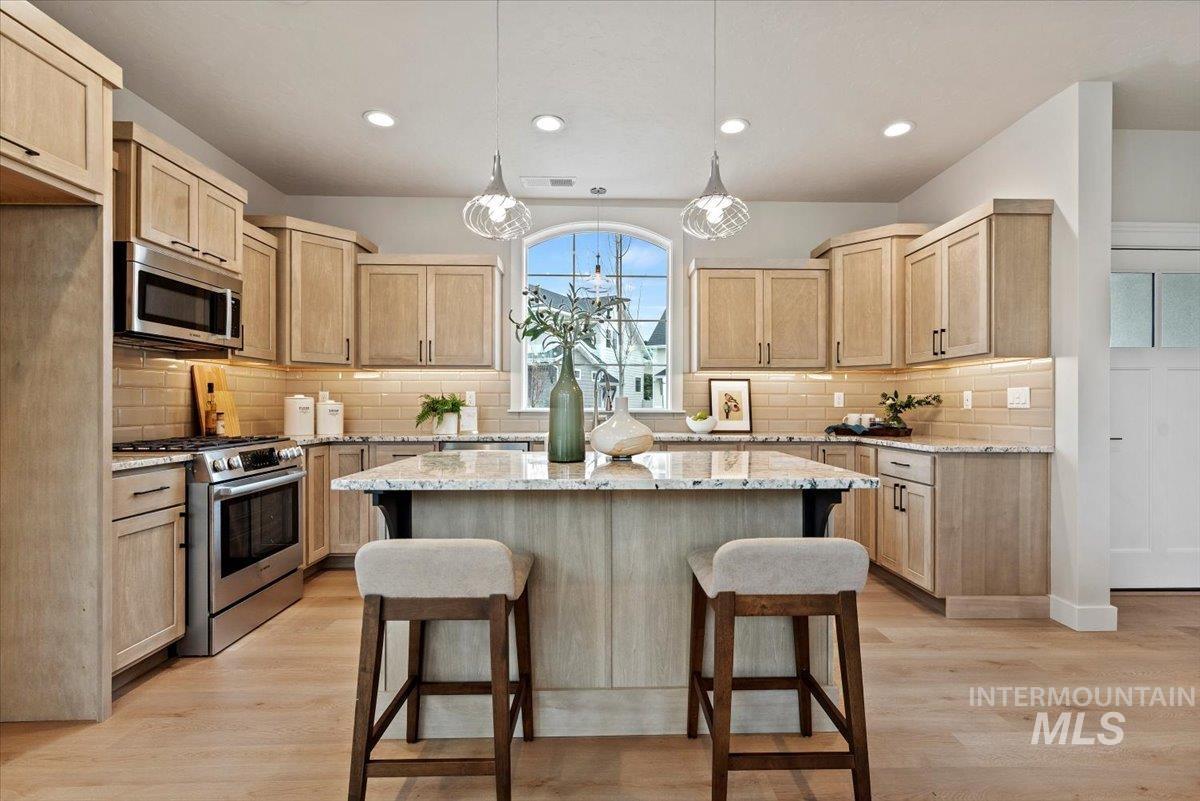 Kitchen with light brown cabinets, hanging light fixtures, appliances with stainless steel finishes, a kitchen bar, and light stone counters
