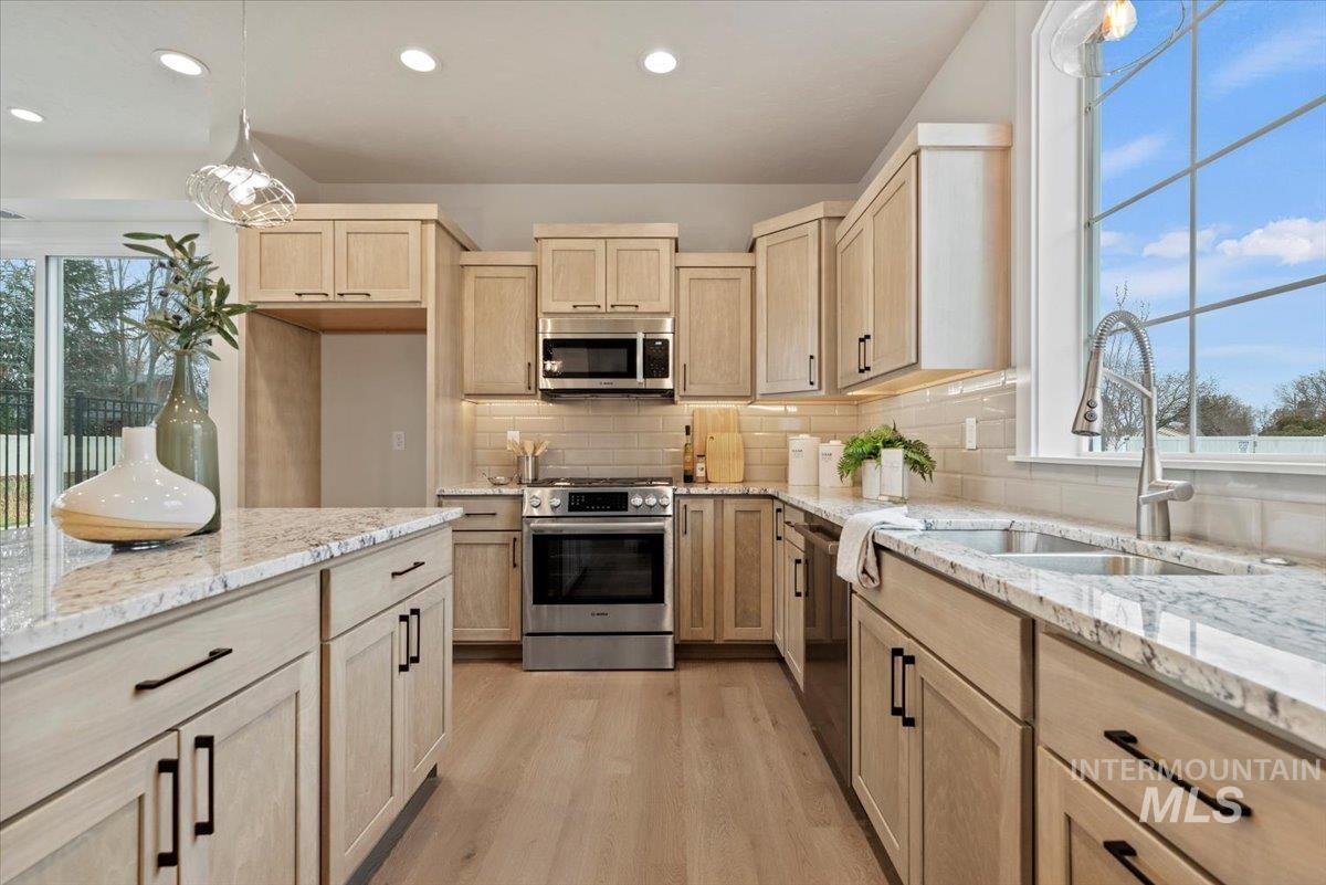 Kitchen with light brown cabinets, pendant lighting, appliances with stainless steel finishes, light stone countertops, and light wood-style floors