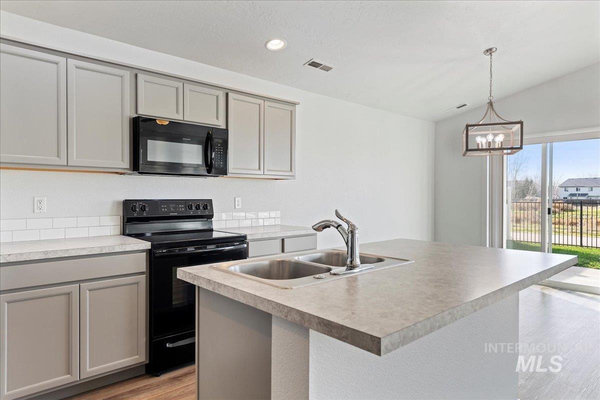 Kitchen featuring black appliances, gray cabinetry, a kitchen island with sink, light countertops, and hanging light fixtures