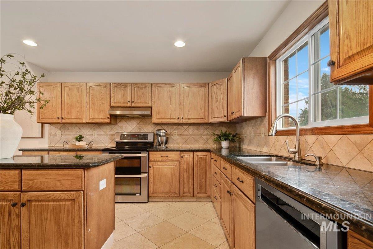 Kitchen with appliances with stainless steel finishes, dark stone counters, decorative backsplash, light tile patterned floors, and under cabinet range hood