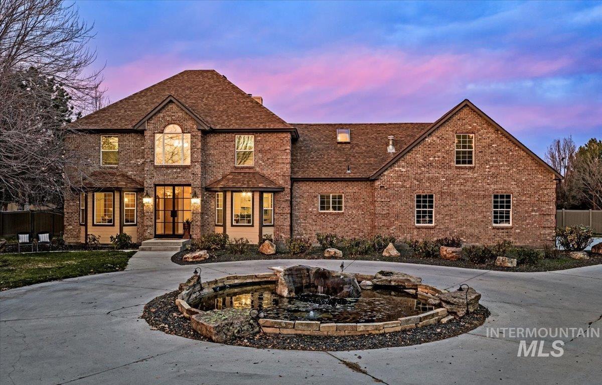 View of front of house with brick siding, curved driveway, a small pond, and roof with shingles