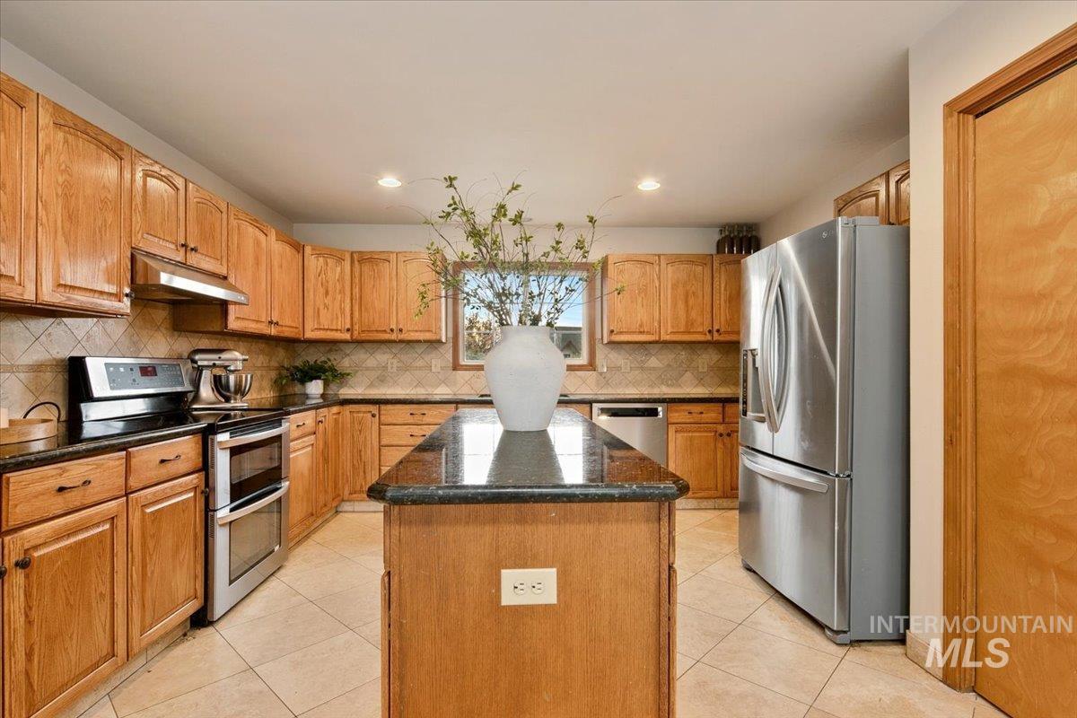 Kitchen with appliances with stainless steel finishes, dark stone countertops, light tile patterned floors, under cabinet range hood, and a kitchen island