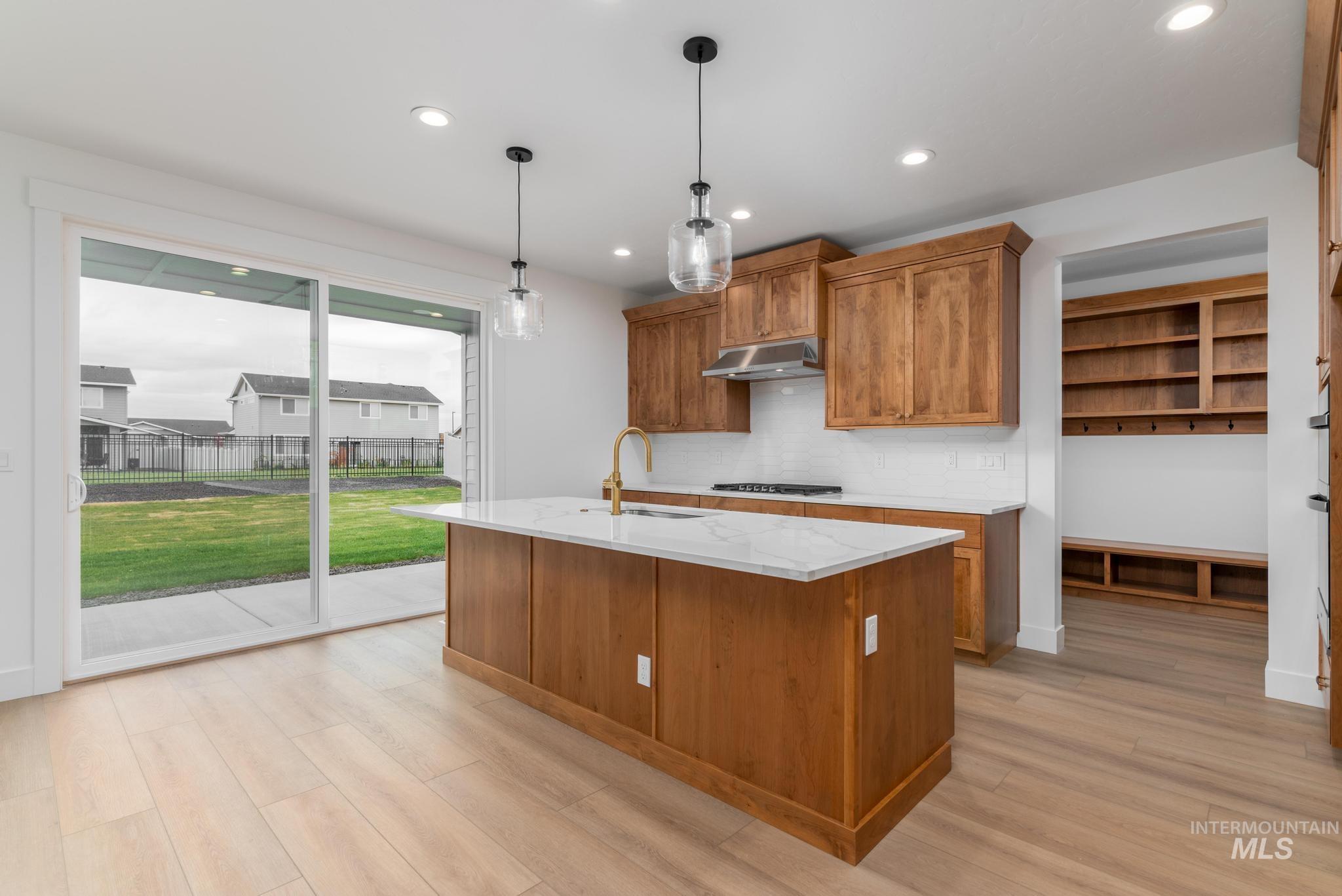 Kitchen featuring brown cabinets, hanging light fixtures, light stone countertops, backsplash, and an island with sink