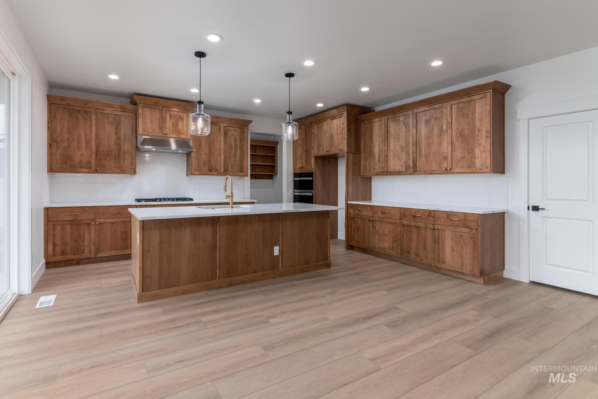 Kitchen with brown cabinetry, decorative light fixtures, light wood-style flooring, a kitchen island with sink, and recessed lighting
