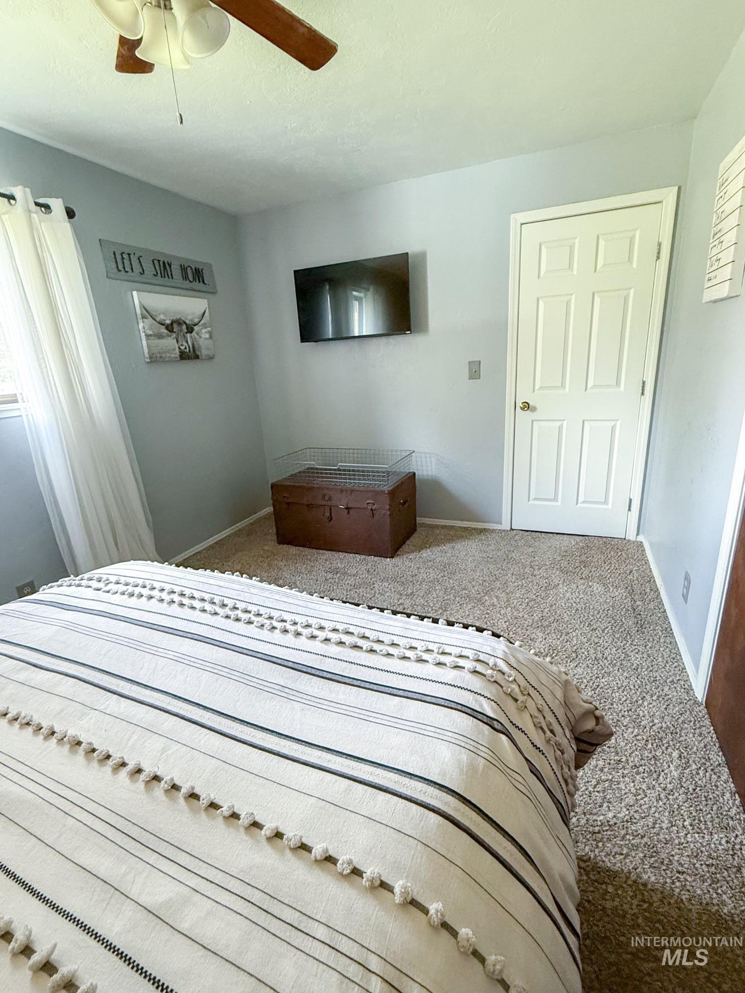 Bedroom featuring light gray walls, textured carpeting, a white paneled door, and a ceiling fan with wood-finish blades