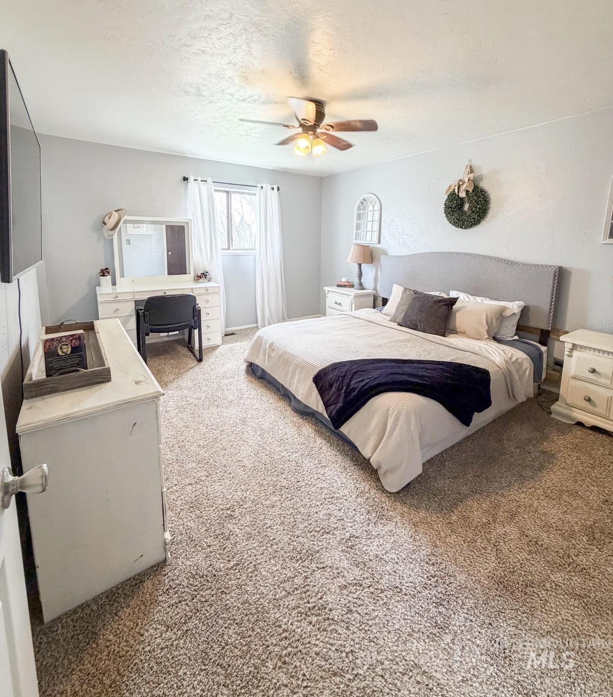 Carpeted bedroom featuring a window with white drapes, a ceiling fan with integrated lighting, and light gray wall paint