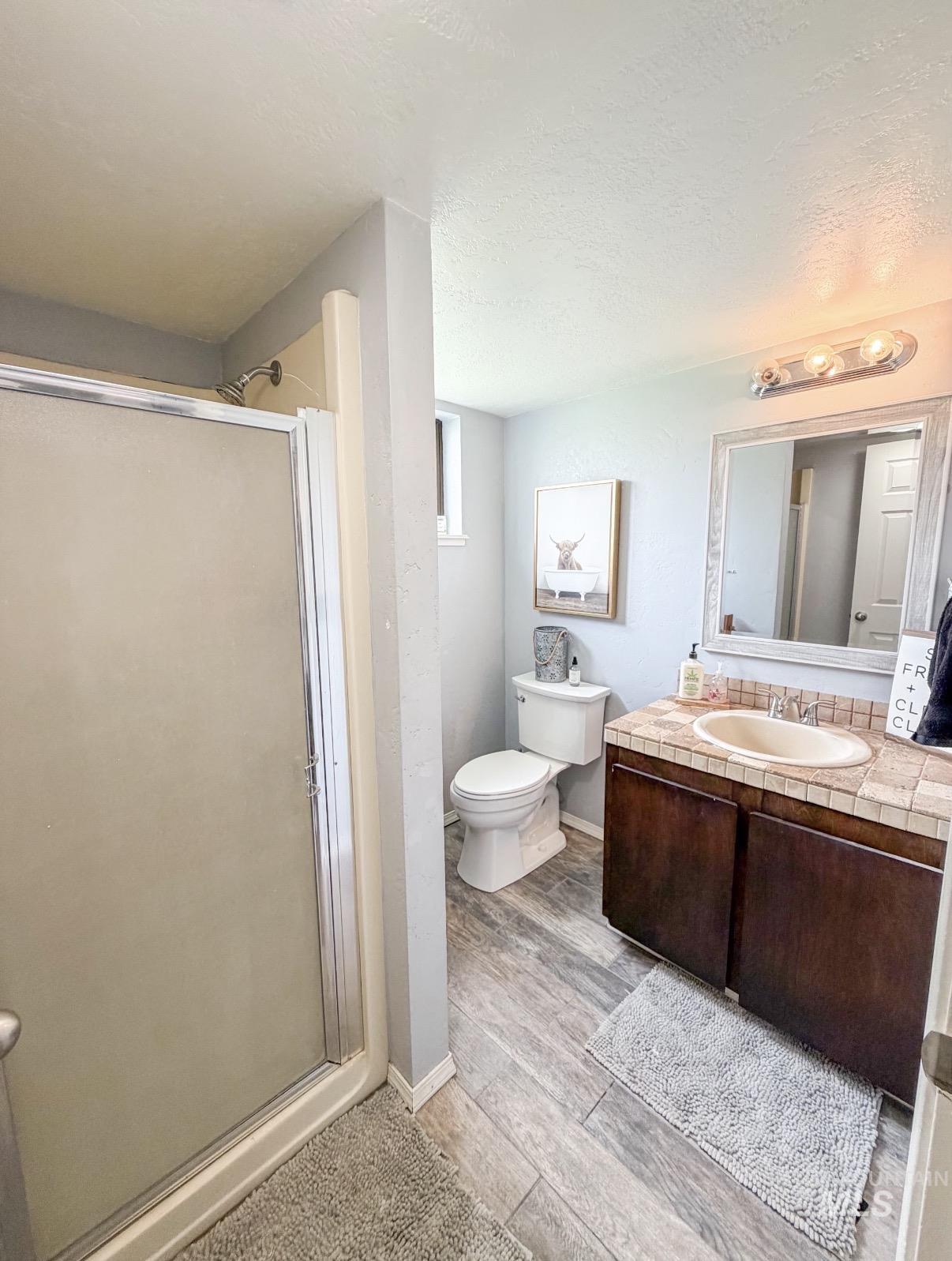 Bathroom featuring a walk-in shower with frosted glass enclosure, single vanity with tile countertop and integrated sink, dark wood-finish cabinetry, wood-look flooring, and recessed window