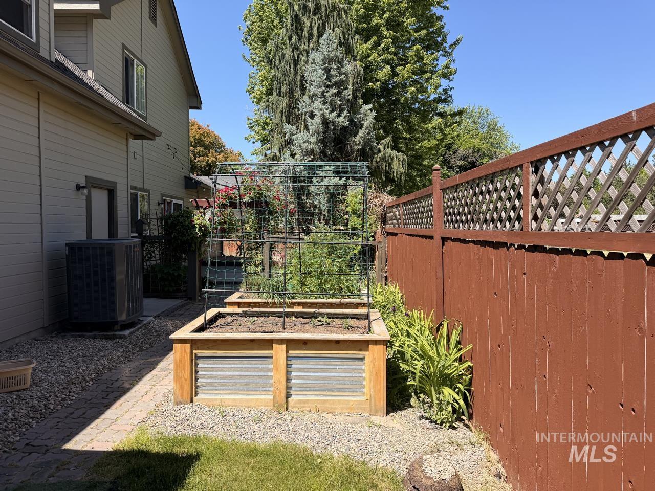 Raised garden beds with integrated trellis, brick paver pathway, gravel landscaping, side yard access, and a wood privacy fence