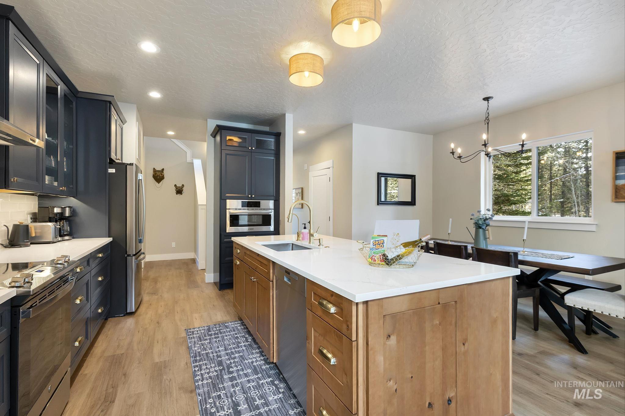Kitchen featuring glass fronted cabinets, a textured ceiling, light wood-style floors, a center island with sink, and stainless steel appliances