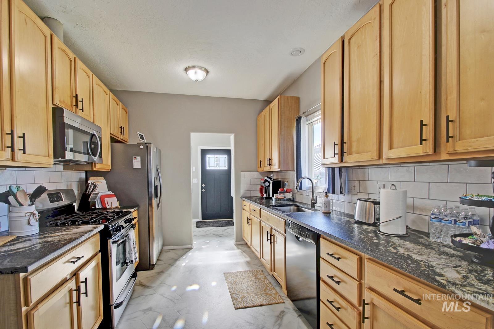 Kitchen featuring stainless steel appliances, decorative backsplash, light marble finish floors, light brown cabinetry, and dark stone countertops