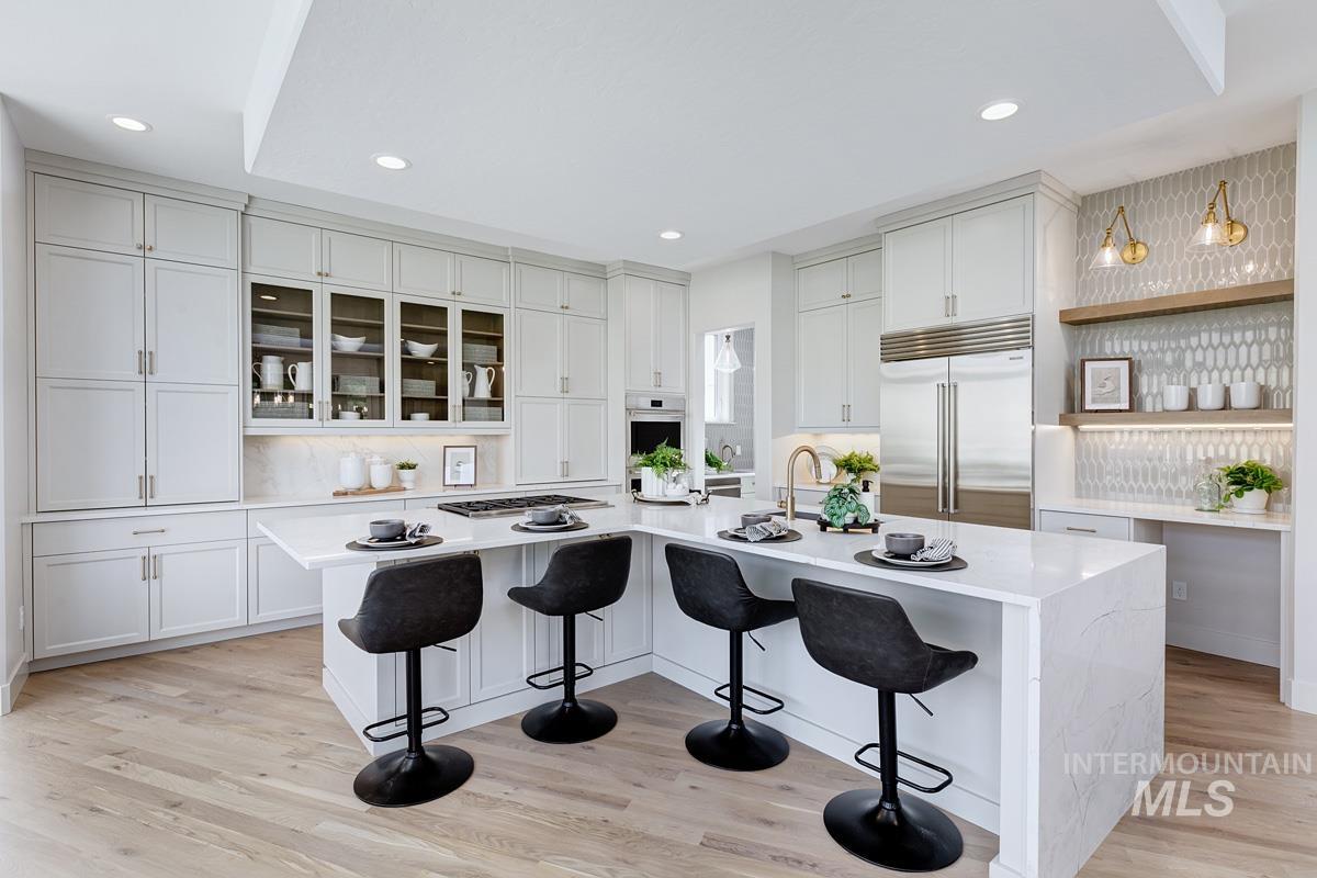 Spacious kitchen featuring a large central island with seating, extensive light-toned cabinetry, integrated stainless steel appliances, wood-finish flooring, and a decorative tiled backsplash with open shelving