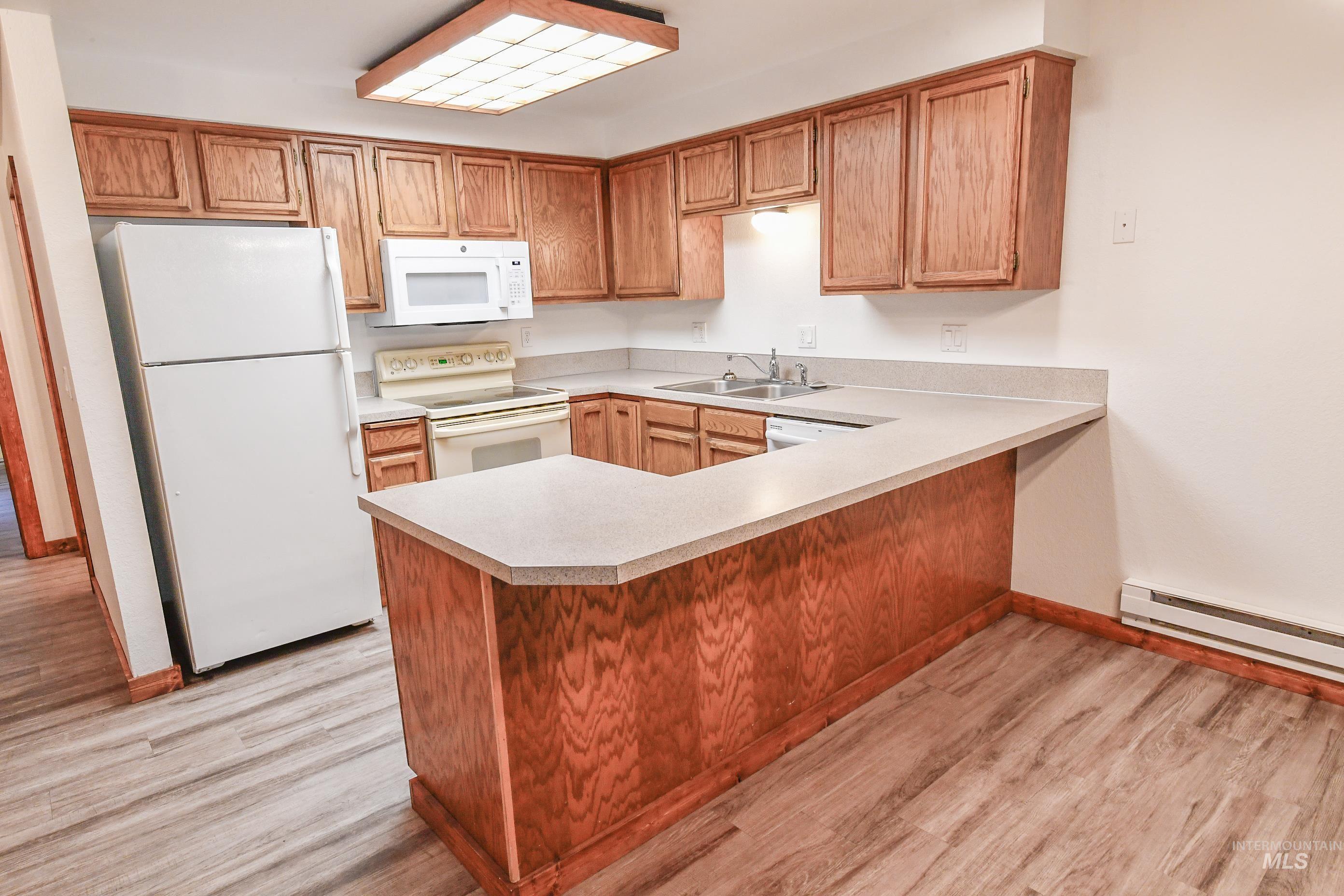 Kitchen featuring white appliances, light countertops, light wood-style floors, baseboard heating, and a peninsula